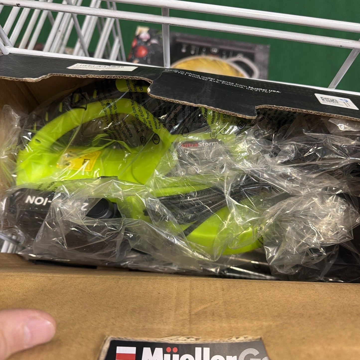 Partially opened box showing a bright green MuellerGo cordless power tool inside, wrapped in clear protective plastic inside a cardboard packaging on a metal store shelf.