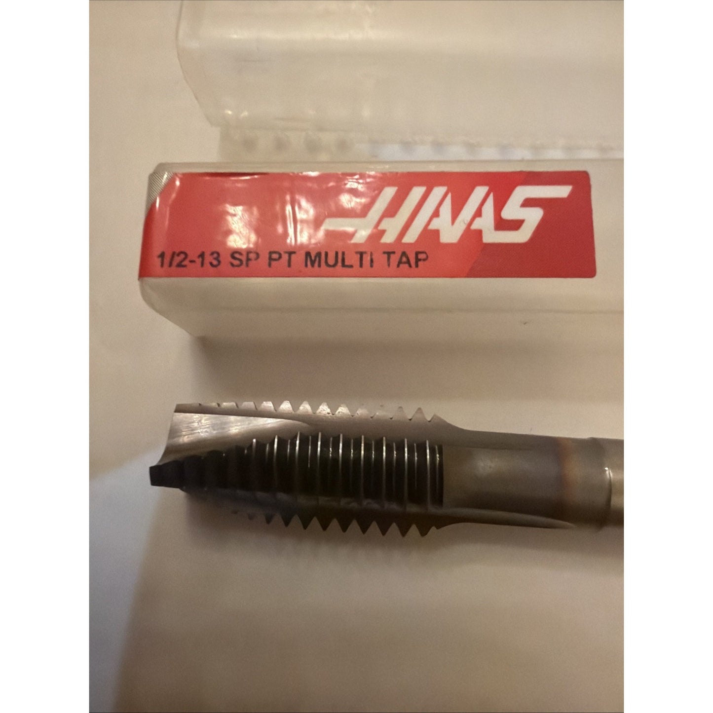 Metal threading tap with spiral-cut threads and a straight shank, positioned below a translucent plastic case featuring a red label with white text on a white surface.