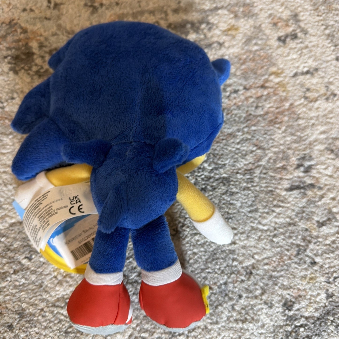 Plush toy of a blue character lying face down on a light-colored textured carpet, showing red shoes, white gloves, and a product tag attached.
