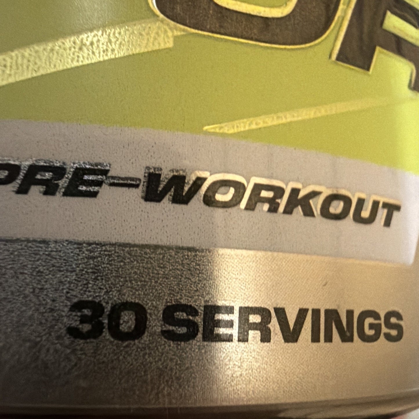 Close-up of a metallic container label displaying "PRE-WORKOUT" and "30 SERVINGS" in bold black text on a green and silver background.