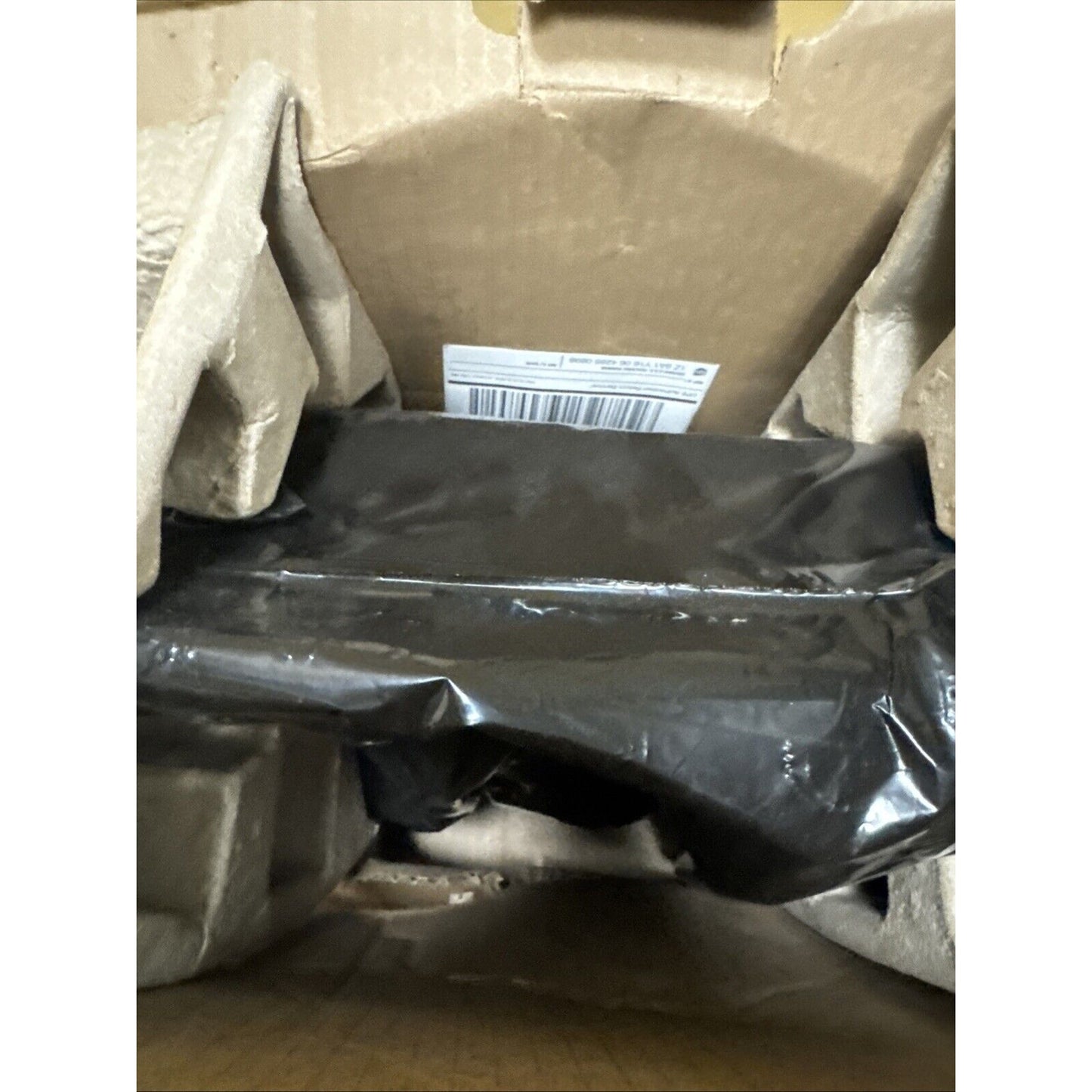 Brown cardboard box interior containing a black plastic-wrapped item secured by molded pulp protective inserts with a visible barcode label inside the box.