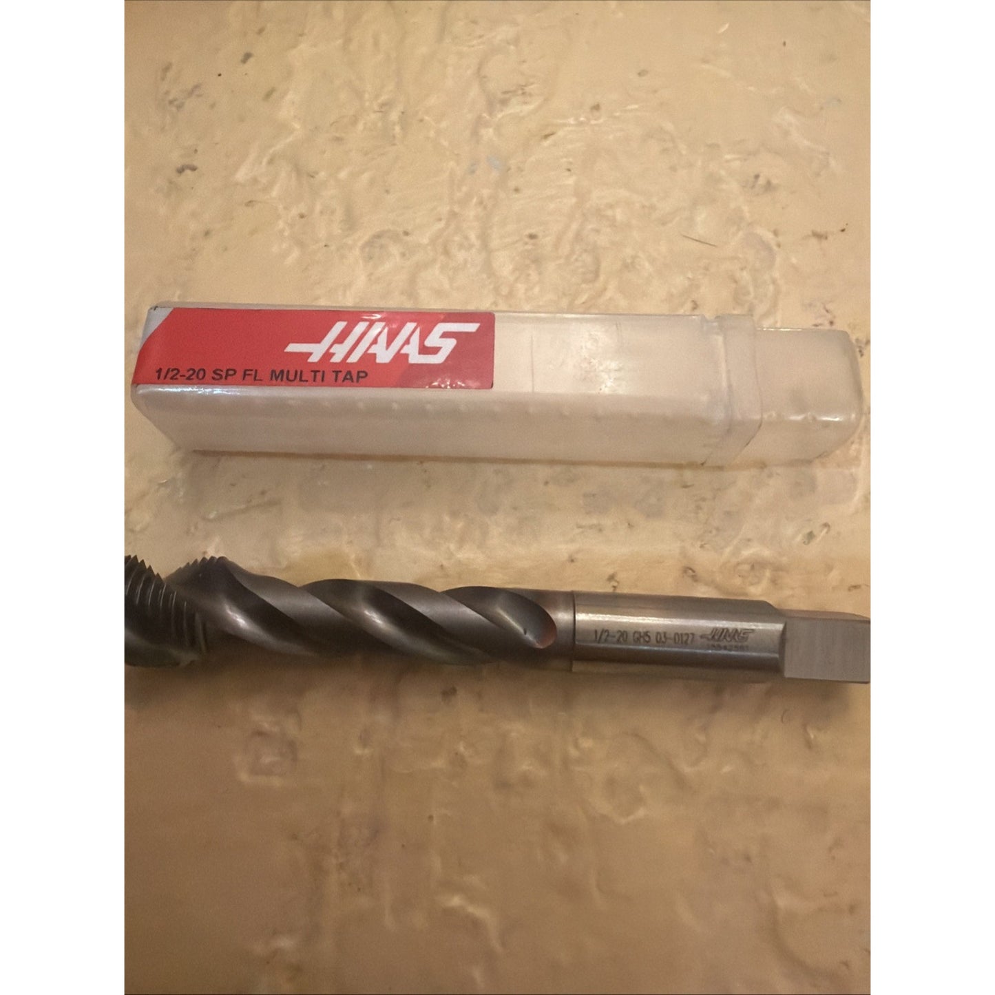 Spiral-fluted metal threading tap with engraved size markings and square shank, lying on a textured beige surface below a clear plastic case featuring a red label with white text.