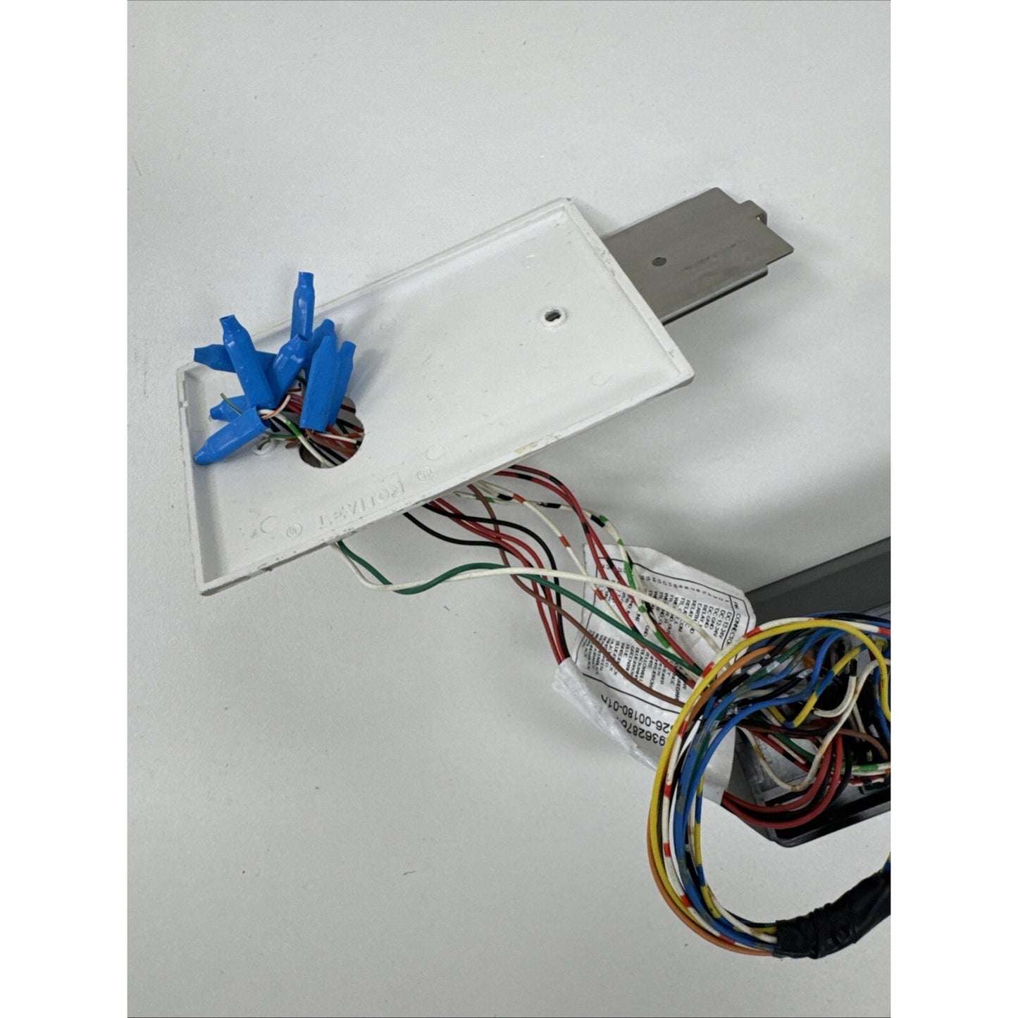 White rectangular plastic panel with multiple blue wire connectors attached to exposed multi-colored electrical wires, connected to a gray metal bracket on a white surface.