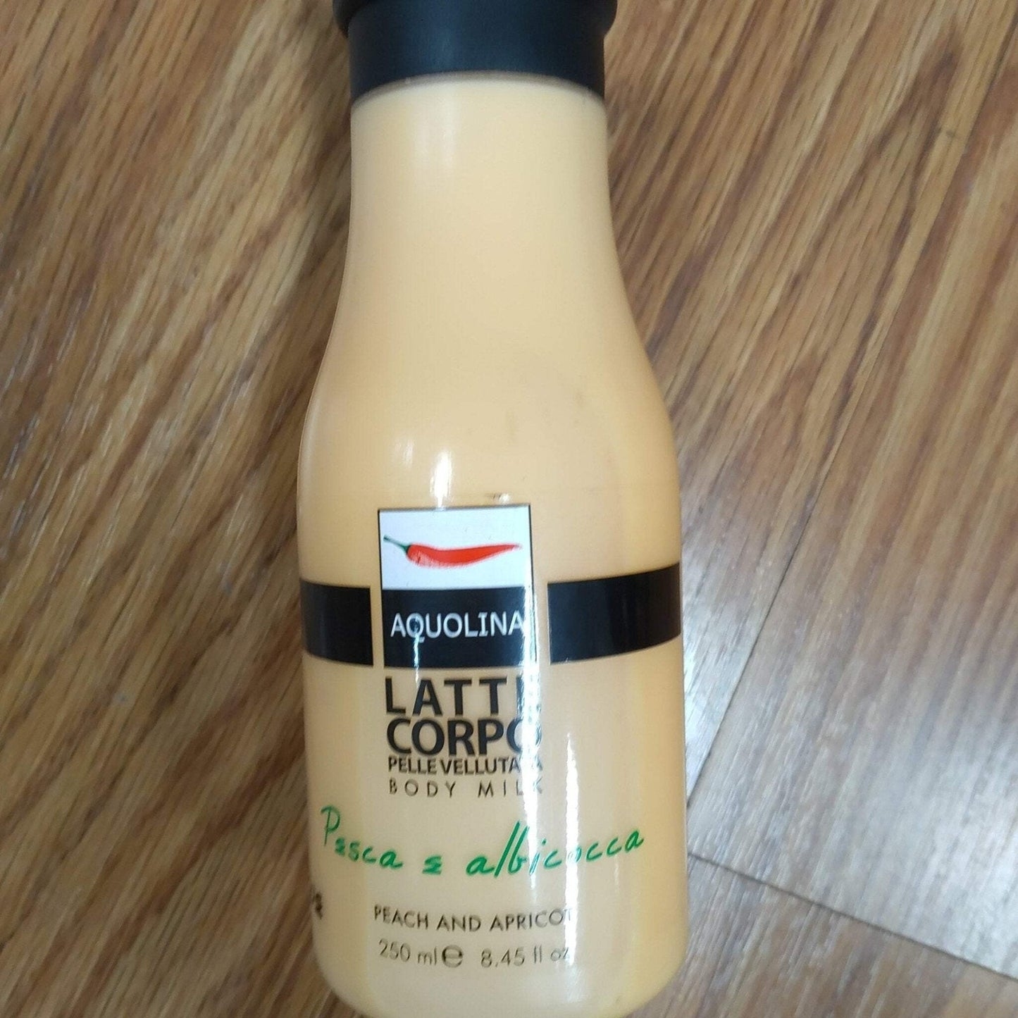 Cream-colored plastic bottle of Aquolina body milk with black cap, labeled in Italian and English indicating peach and apricot scent, placed on a wooden surface.