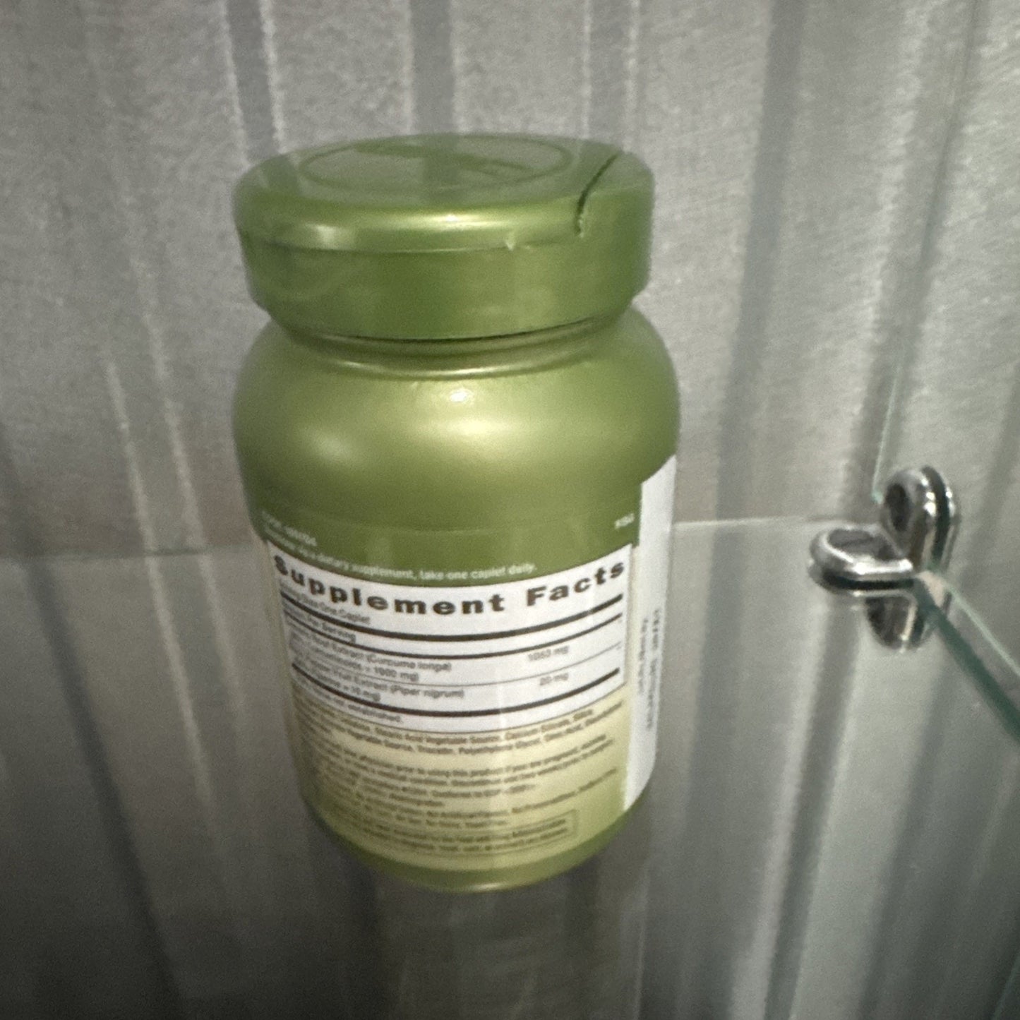 Green plastic bottle with a child-resistant cap displaying a white Supplement Facts label, placed on a glass shelf with a blurry textured gray background.