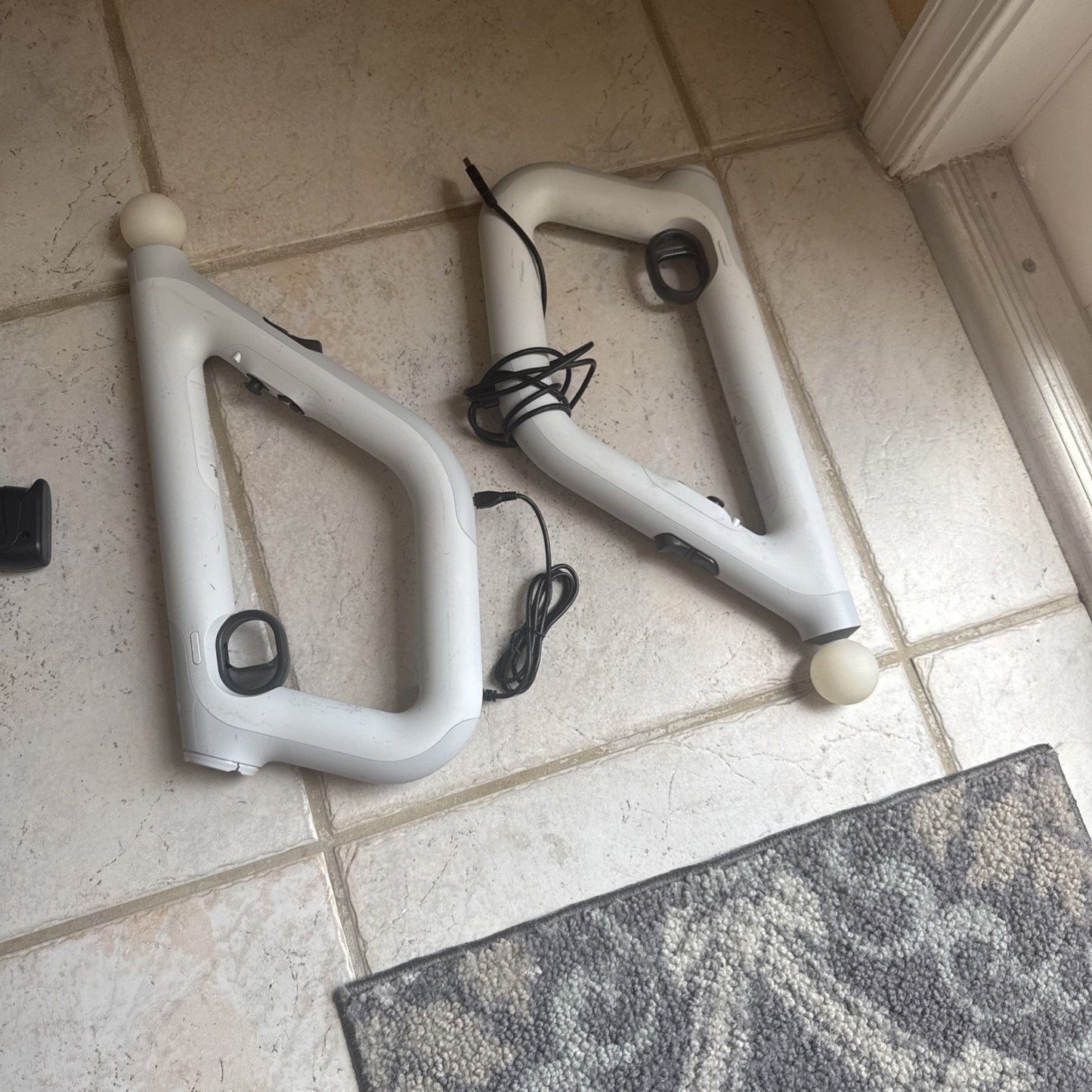 Two white PlayStation VR Aim Controllers with attached cables on tiled floor near rug edge