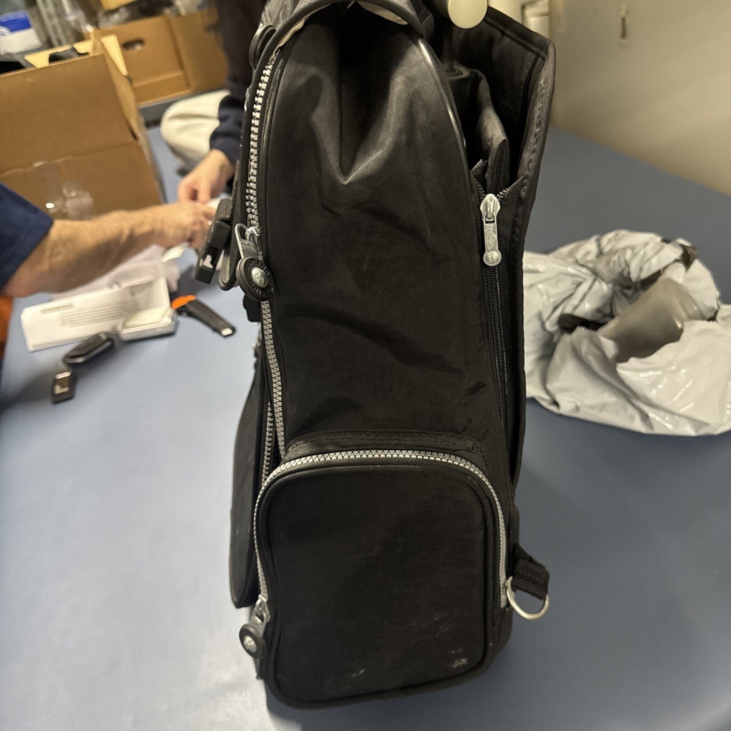 Side view of black Kipling Gaze large printed rolling backpack with retractable handle and front zippered pocket on blue surface