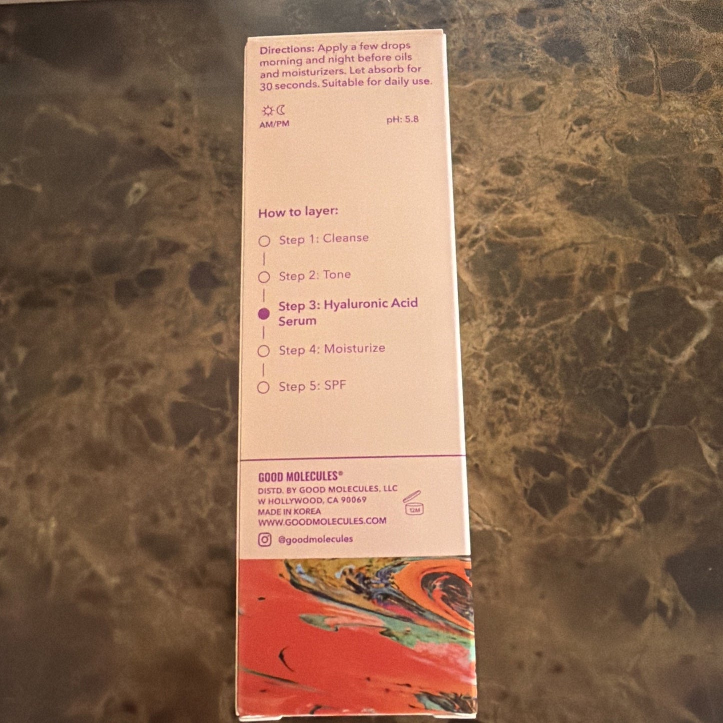 Side panel of a skincare product box displaying usage directions, layering steps including cleanse, tone, hyaluronic acid serum, moisturizer, and SPF, with contact and manufacturing details at the bottom.