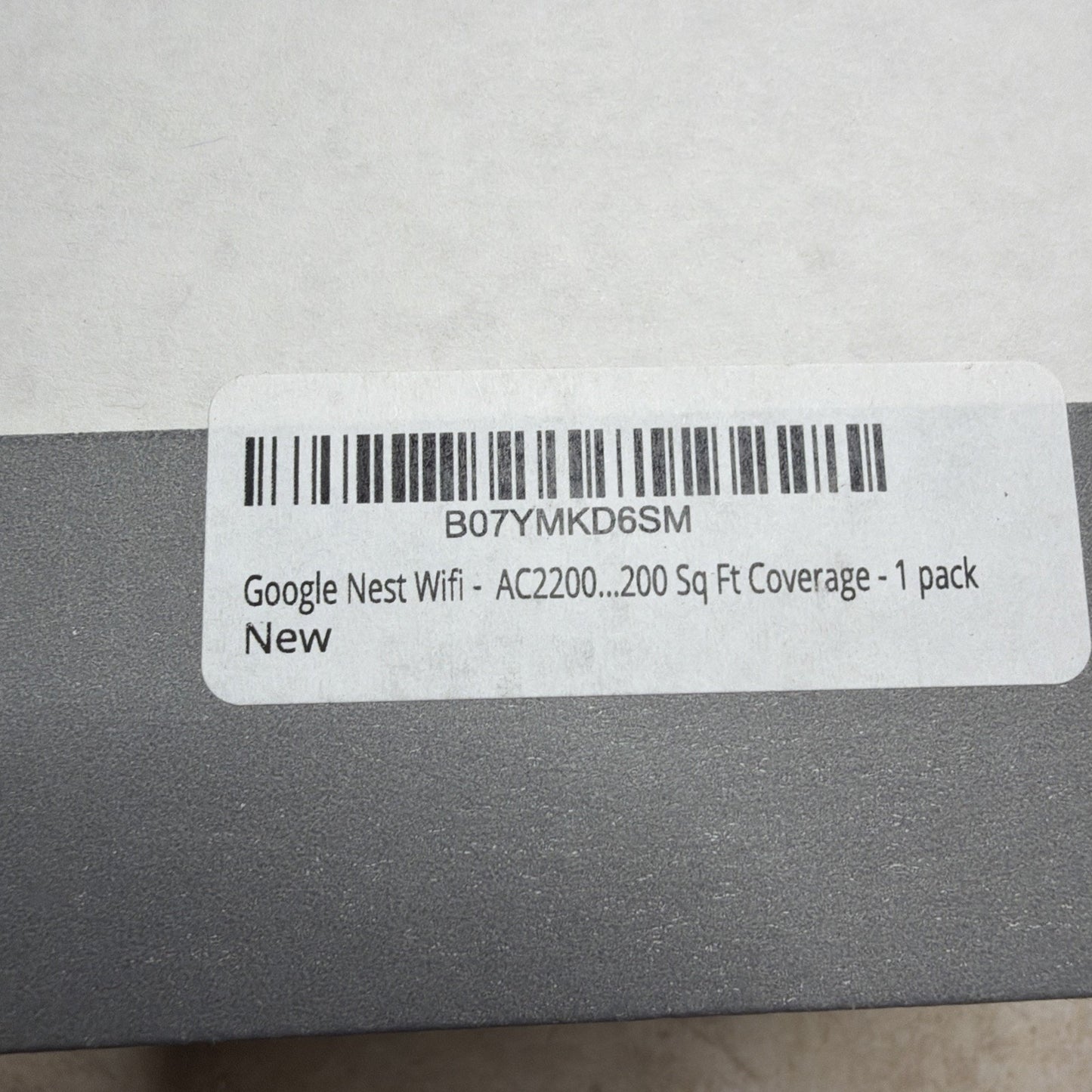 Close-up of a white barcode label on a gray surface showing product information for a single pack of Google Nest Wifi with AC2200 and coverage of 200 square feet, marked as new.
