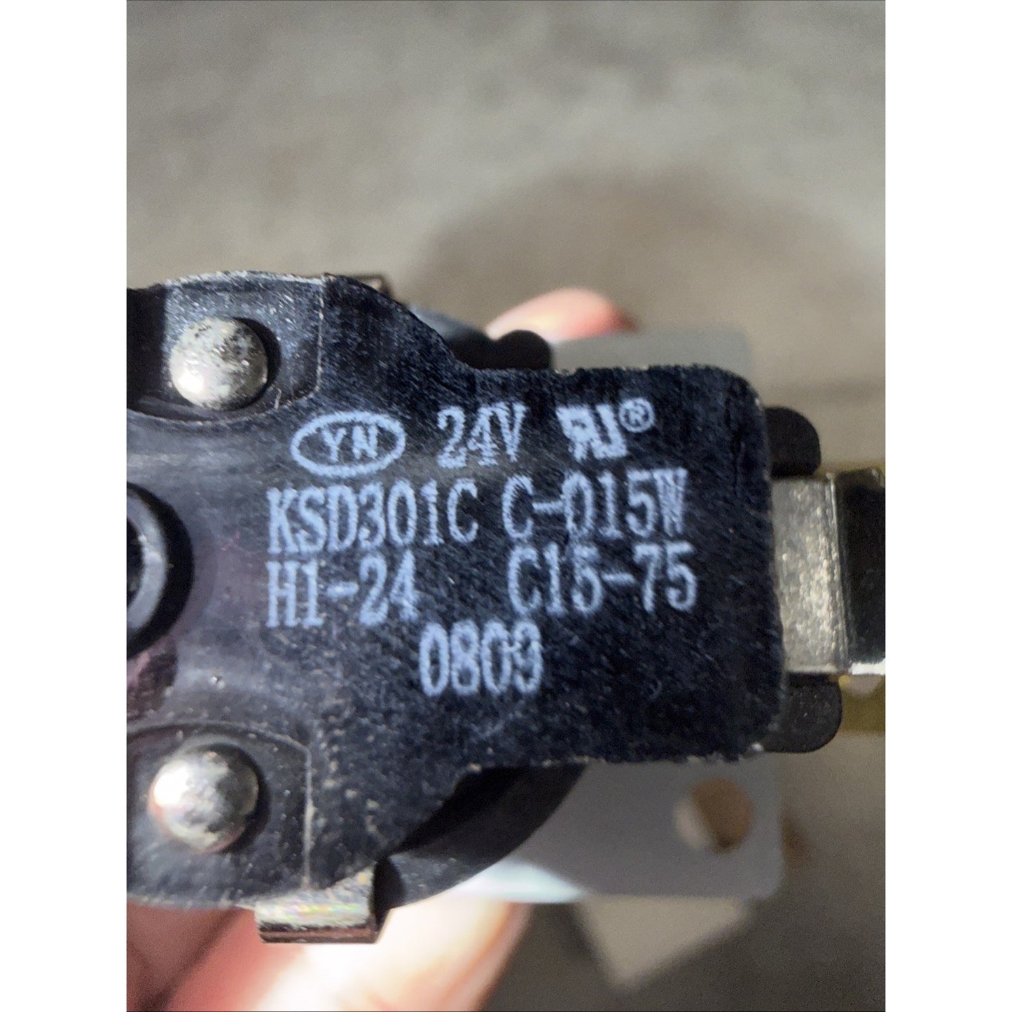 Close-up of a black electrical component with white printed specifications including "24V," "KSD301C," and other alphanumeric codes, held by a hand against a blurred background.