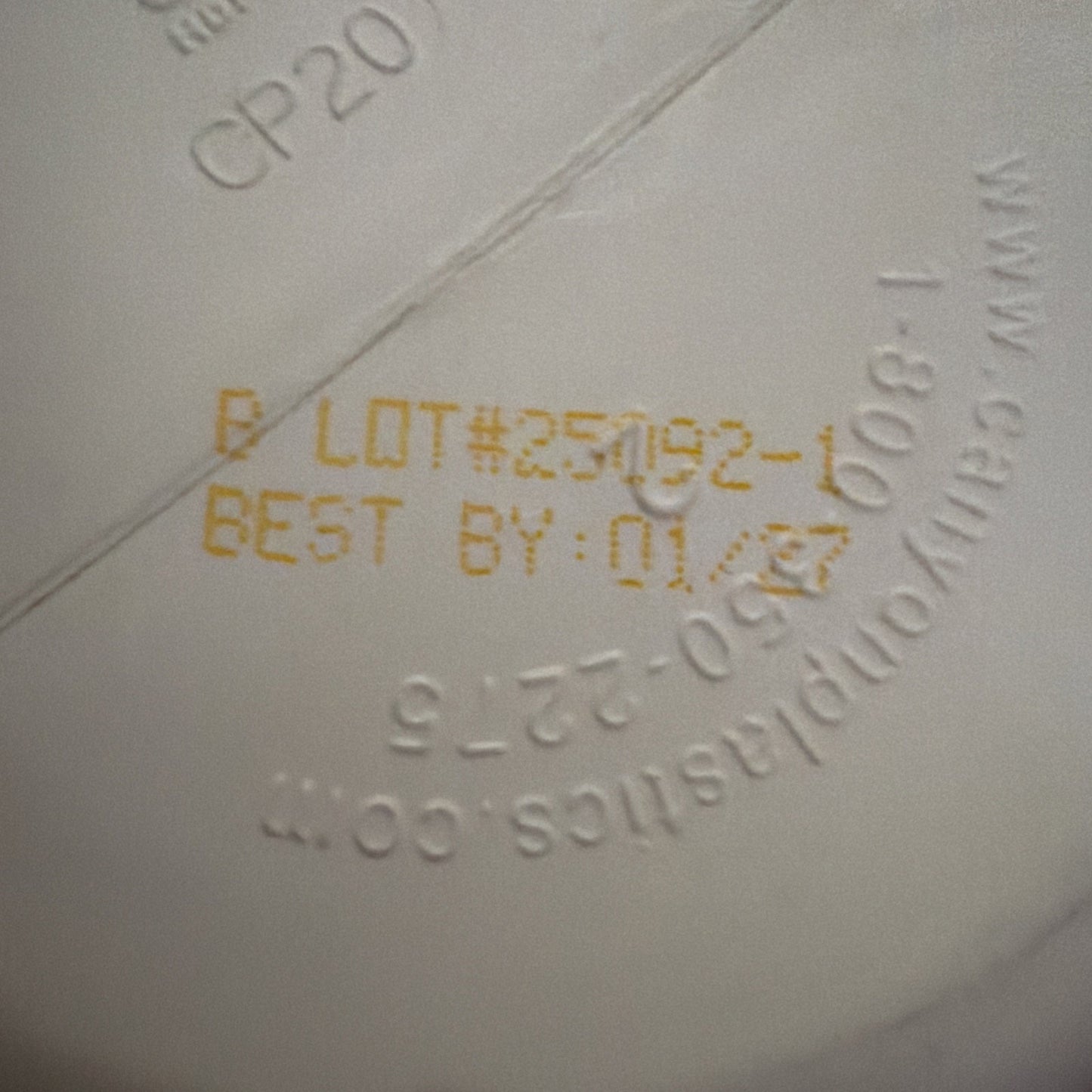 Close-up of a white container bottom showing an embossed website URL and printed yellow batch number "25092-1" with best-by date "01/27" on a smooth surface.