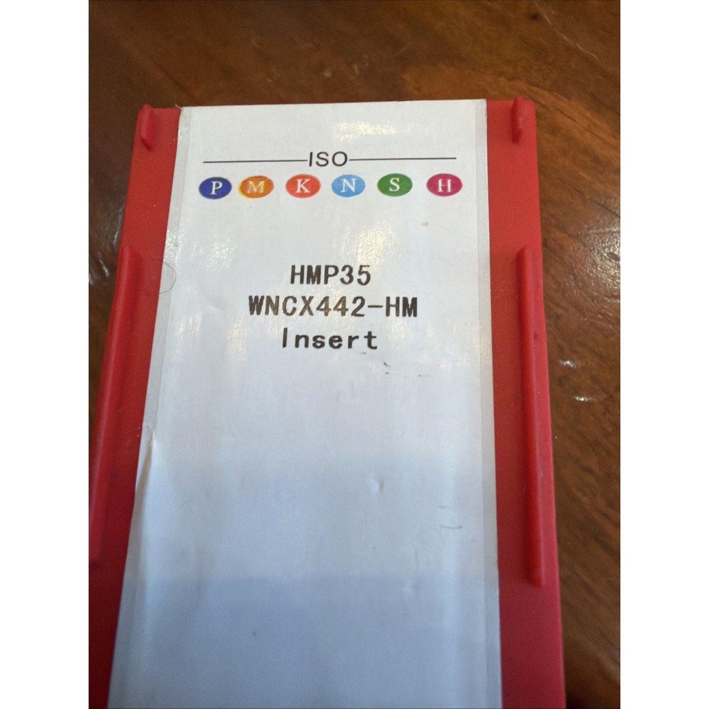 Red plastic container with a white label displaying colored ISO classification symbols and text reading "HMP35 WNCX442-HM Insert" on a wooden surface.