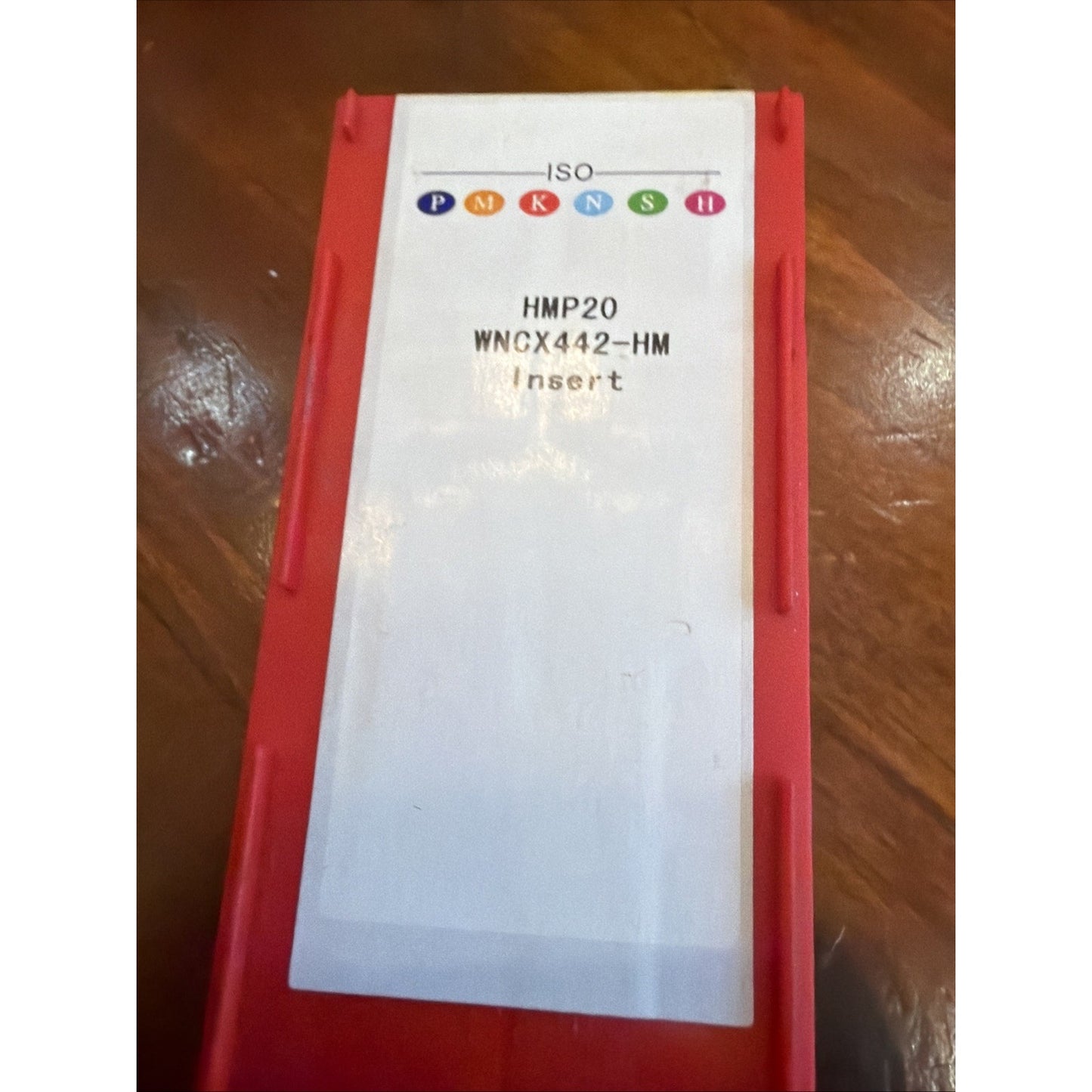 Rectangular red plastic container with a white label displaying colored ISO standard symbols and text reading "HMP20 WNCX442-HM Insert" on a wooden surface.