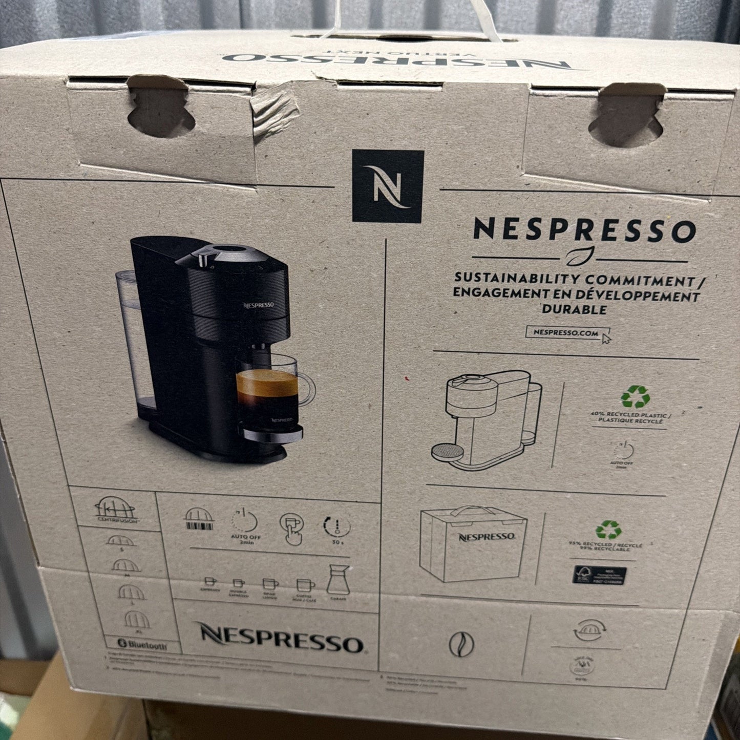 Cardboard packaging for a Nespresso coffee machine featuring a photo of the black machine with a cup of espresso, icons for device features, and text highlighting sustainability and recyclable materials.