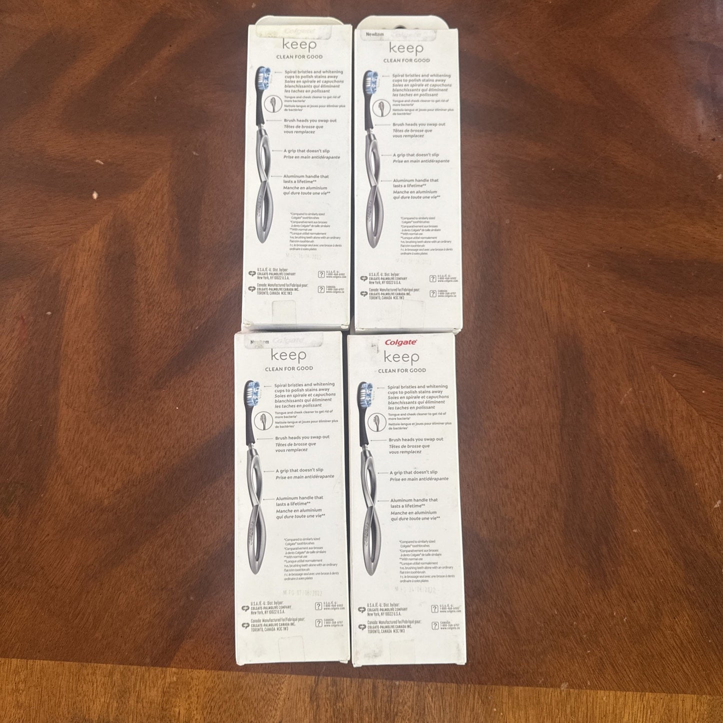 Four white rectangular boxes of Colgate Keep Clean Floss toothbrushes arranged in a 2x2 grid on a wooden surface, showing product information and images of the toothbrush handle and bristles on the back.