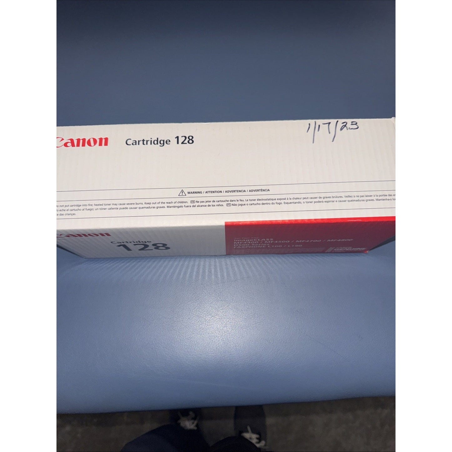 White and red Canon toner cartridge box labeled "Cartridge 128" with a handwritten date on the top right corner, resting on a smooth blue surface.