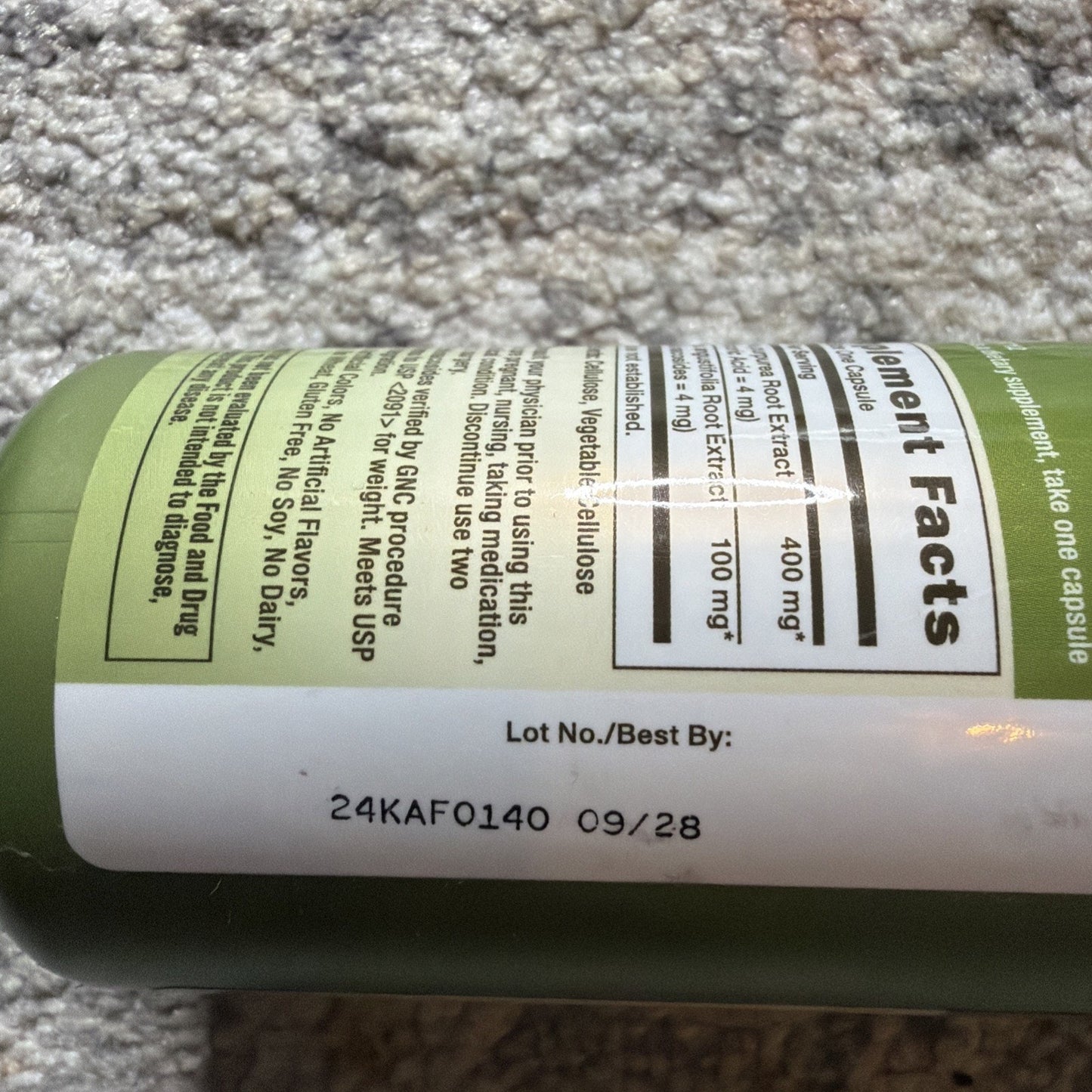 Close-up of a green supplement bottle label showing lot number 24KAF0140 and best by date 09/28, with partially visible ingredient facts and usage instructions.