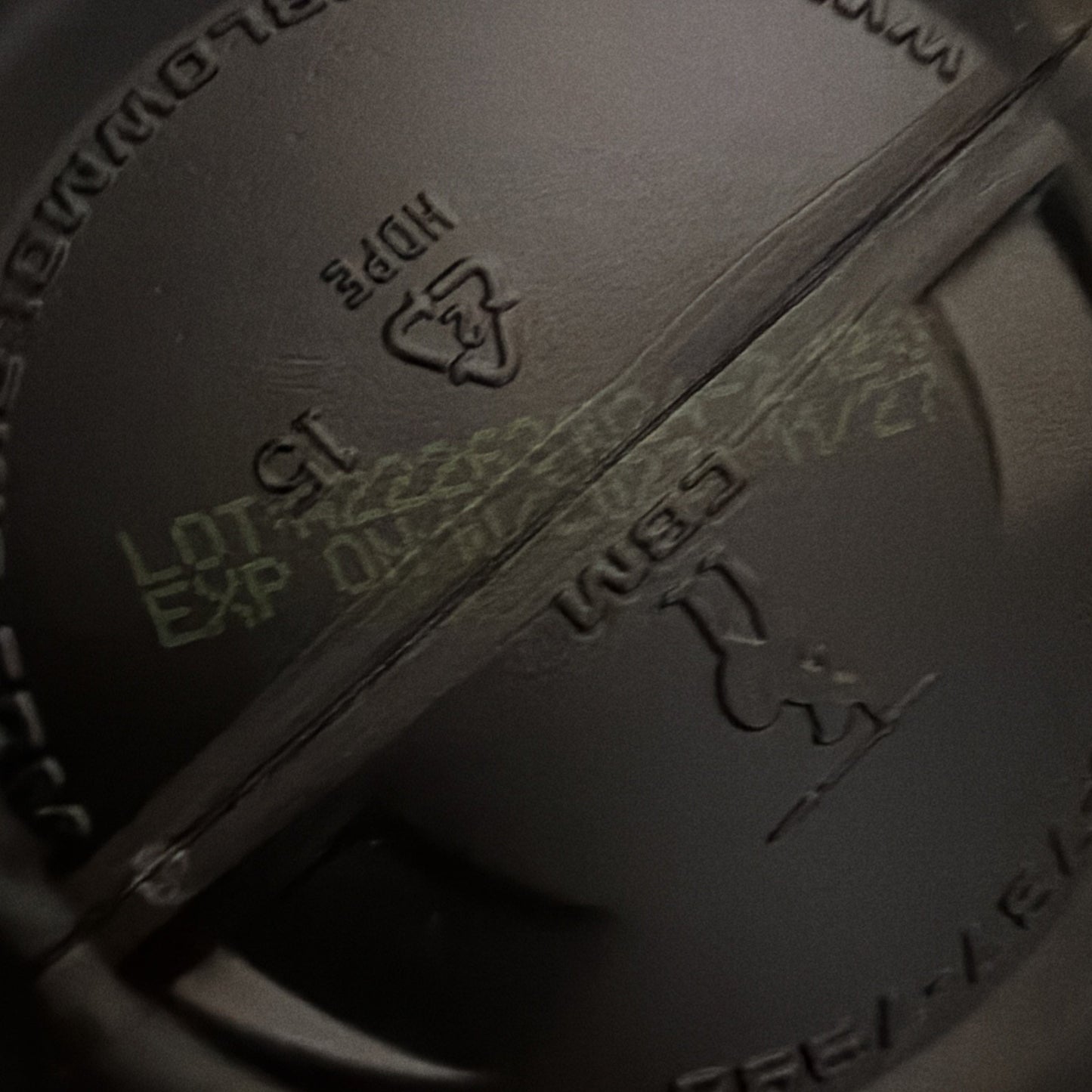 Close-up of pale yellow batch code and expiration date printed on the inside bottom of a dark plastic container lid with molded recycling and material symbols.