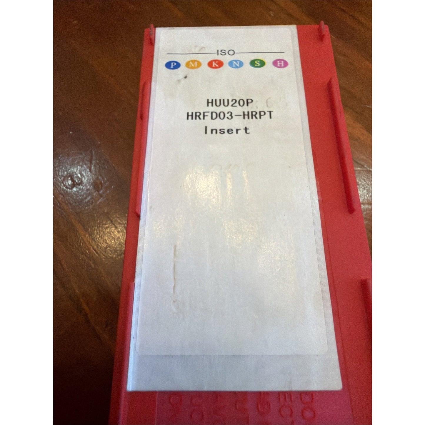 Red plastic storage case with a white label displaying ISO classification symbols in multiple colors and text "HUU20P HRFD03-HRPT Insert," placed on a wooden surface.