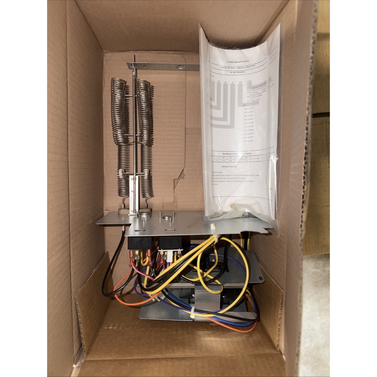 View inside an open cardboard box containing an electrical heating element assembly with metal coils, multiple colored wires, a metal mounting plate, and a plastic sleeve holding printed instructions.