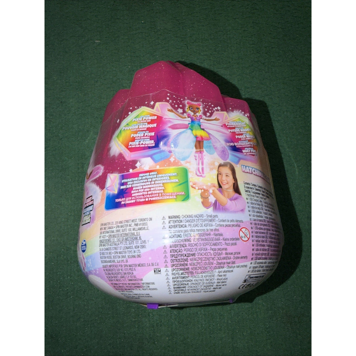 Pink and white toy packaging shaped like an egg, featuring a colorful fairy character illustration, product information, and a photo of a smiling girl holding the toy against a green background.
