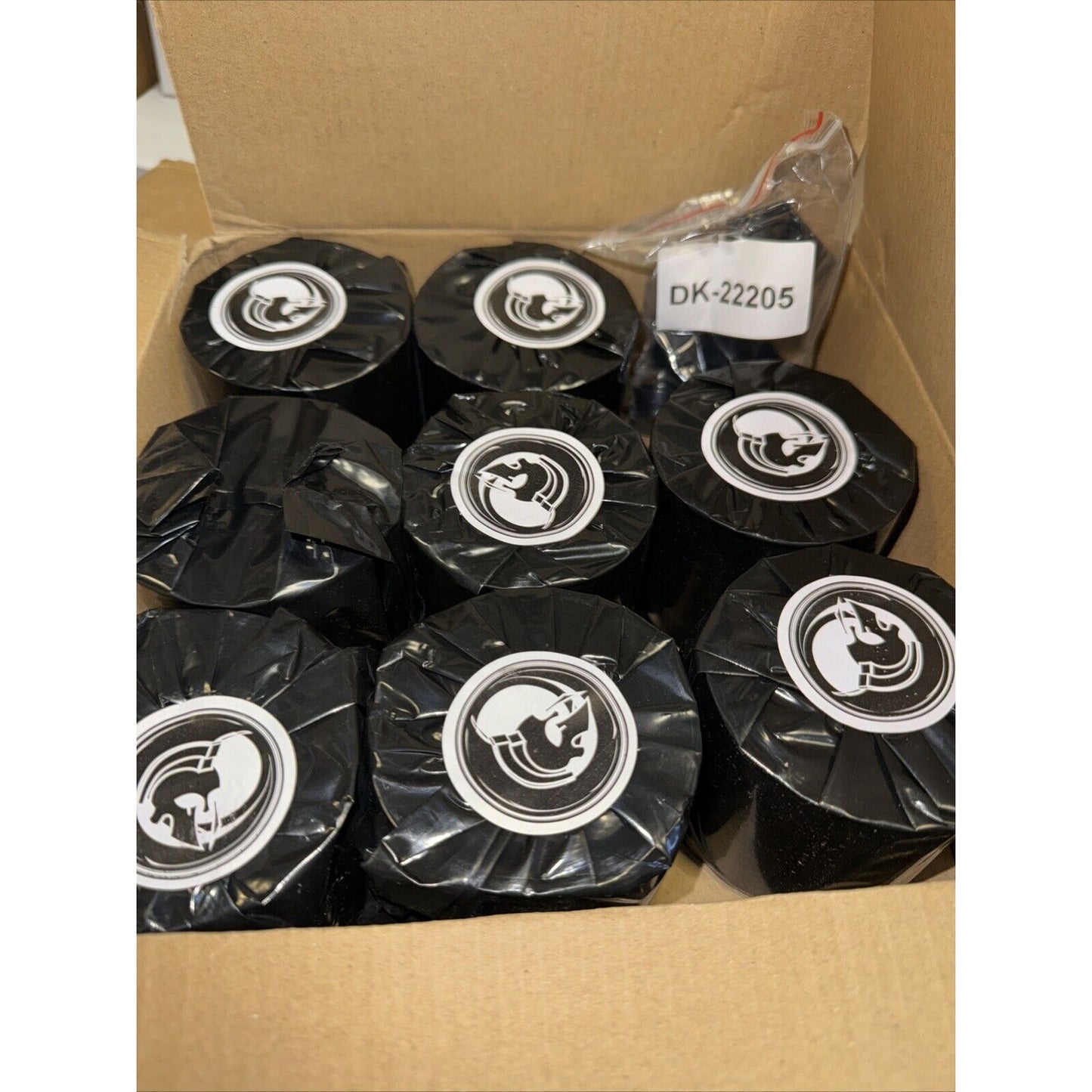 Open cardboard box containing several black rolls wrapped in plastic, each with a round white label featuring a stylized rooster design, alongside a small white tag marked "DK-22205.