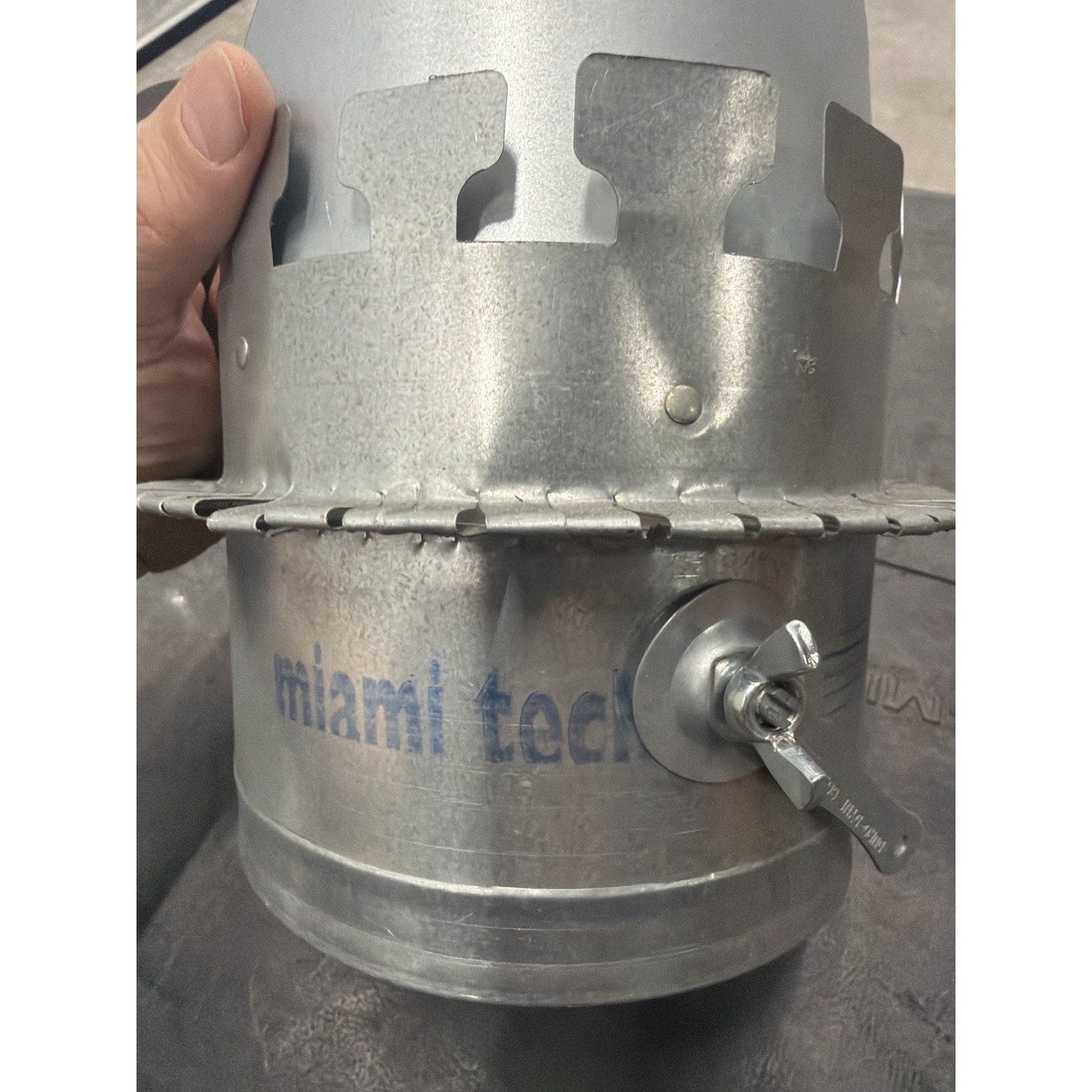 Close-up of a galvanized metal duct component featuring a circular clamp and a small wing nut valve handle, with faint blue text partially reading "miami tech" stamped on the surface.