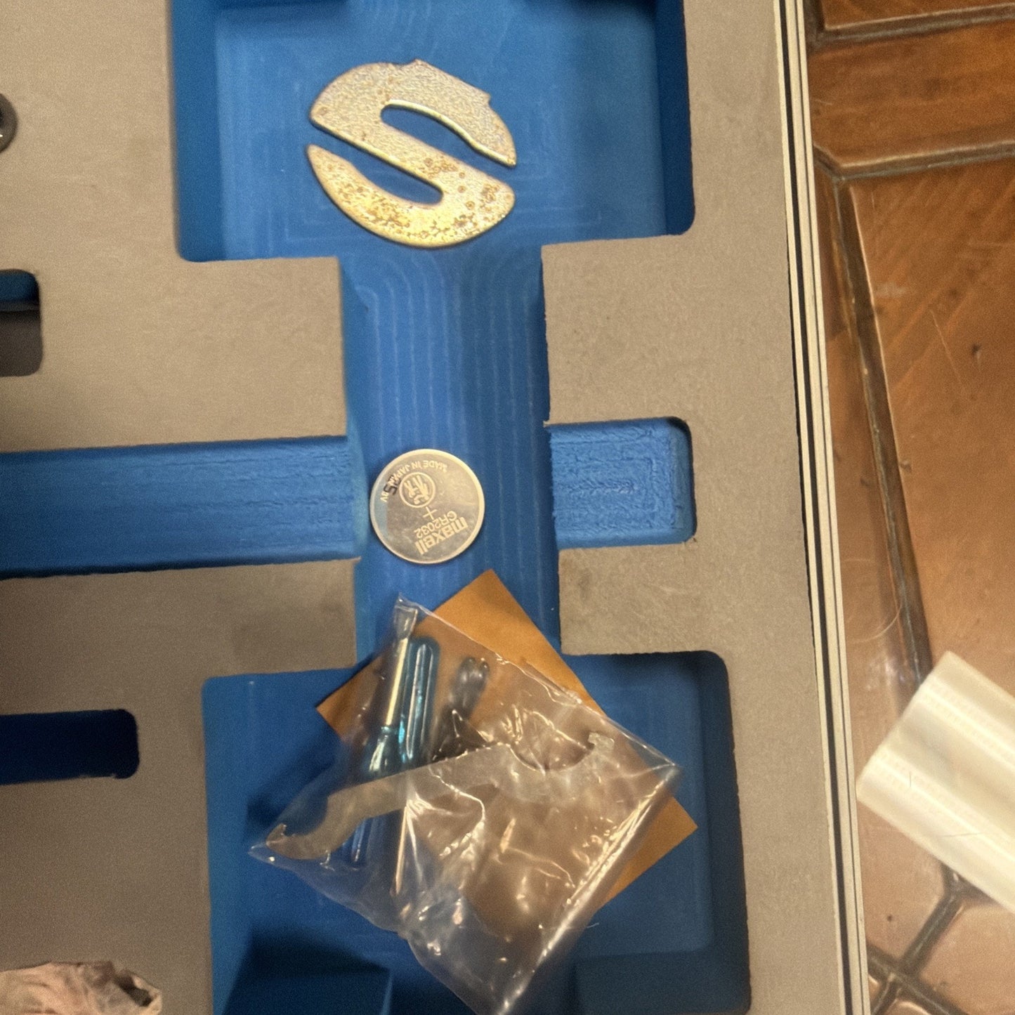 Blue foam insert with several small items including a round silver coin cell battery, a large metallic S-shaped piece, and a small plastic bag containing screws and metal parts, all placed inside a gray case on a wooden floor.
