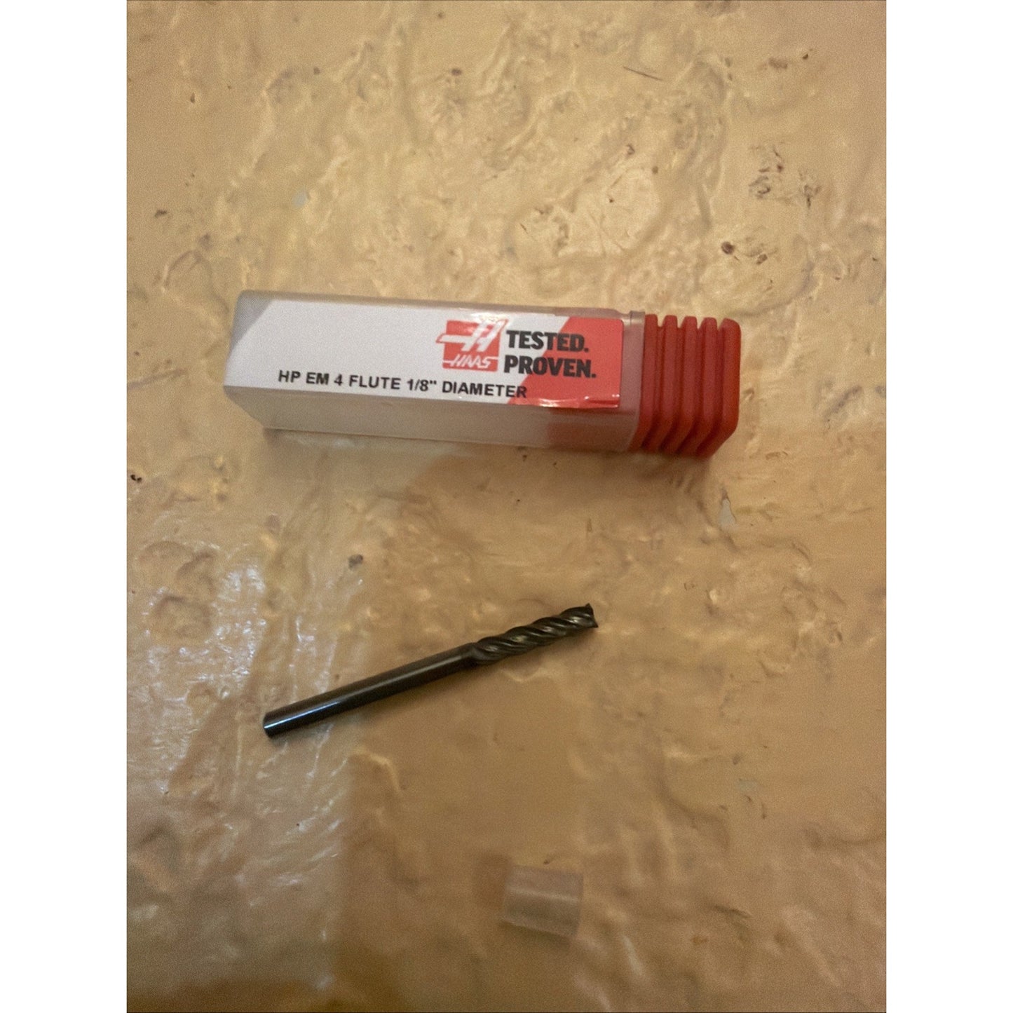 A single dark metal four-flute cutting tool with a small plastic cap lies beneath a rectangular plastic storage case labeled with a red and white Haas logo and text indicating "HP EM 4 FLUTE 1/8" DIAMETER.