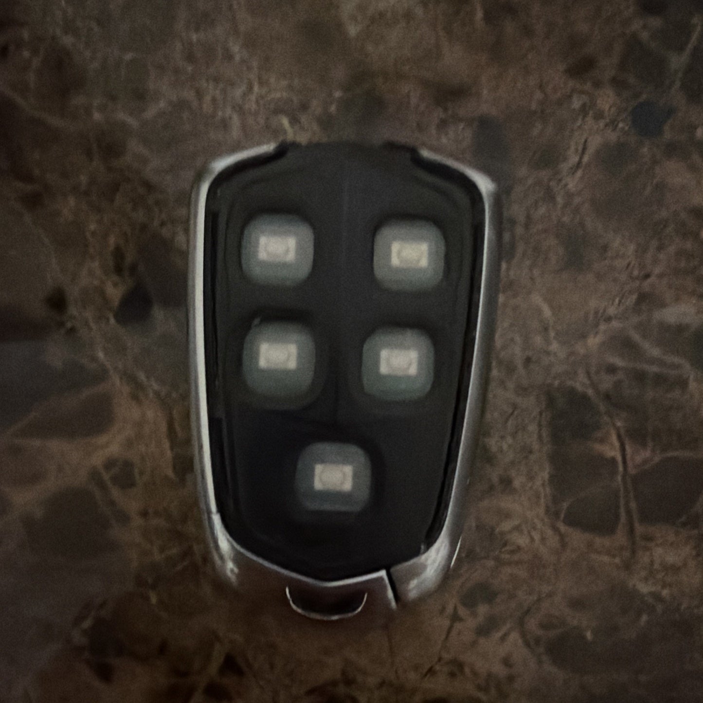 Close-up of a black car key fob with five rubberized buttons set in a chrome-trimmed casing, placed on a dark marbled surface.