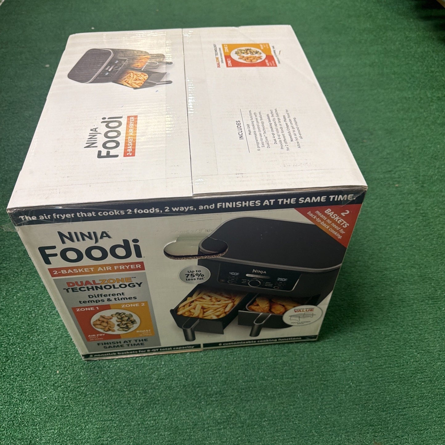 White rectangular box featuring a Ninja Foodi 2-basket air fryer with dual-zone technology, showing images of the appliance cooking fries and other foods simultaneously on a green carpeted surface.