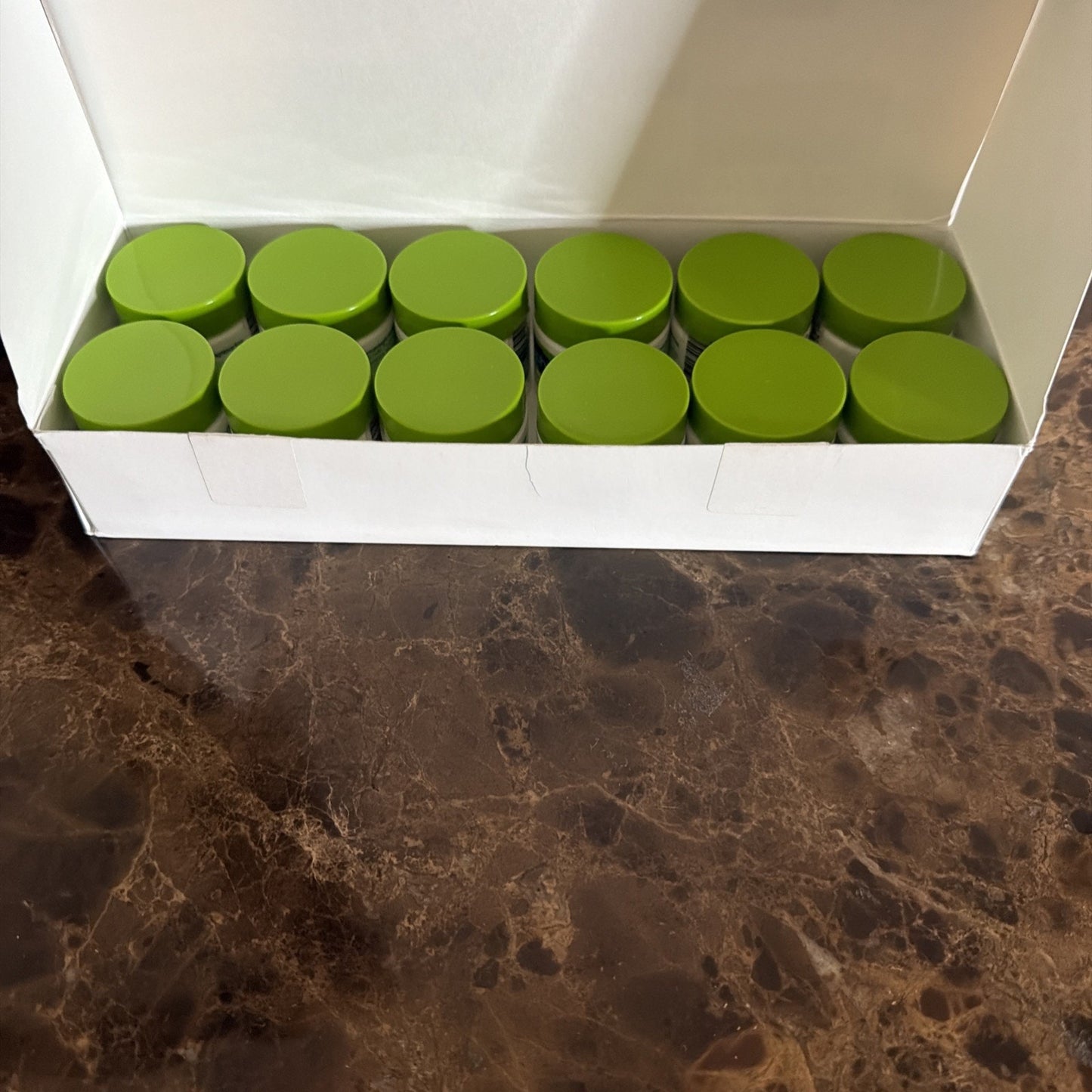 White rectangular box opened on a dark marbled surface containing twelve small containers with green lids arranged in two rows.