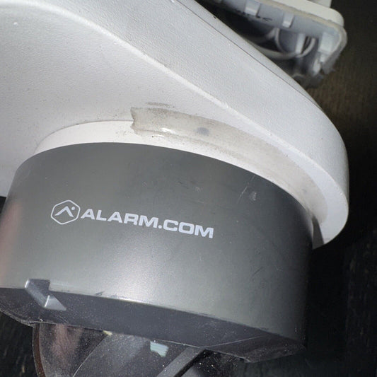Alarm.com surveillance camera close-up showing brand logo on gray body with white mounting base and wiring visible