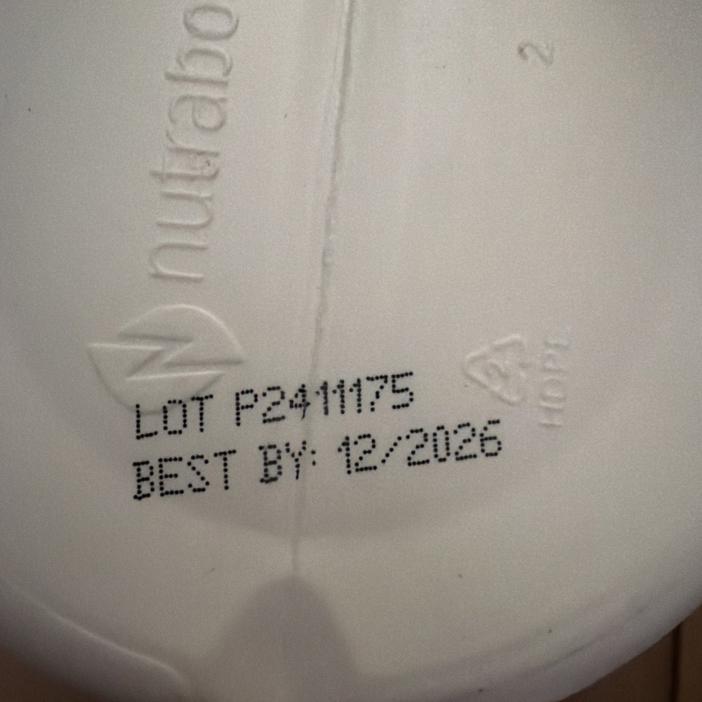 Close-up of a white container bottom with embossed brand name "nutra" and recycling symbol "2 HDPE," showing printed lot number "P2411175" and best-by date "12/2026.