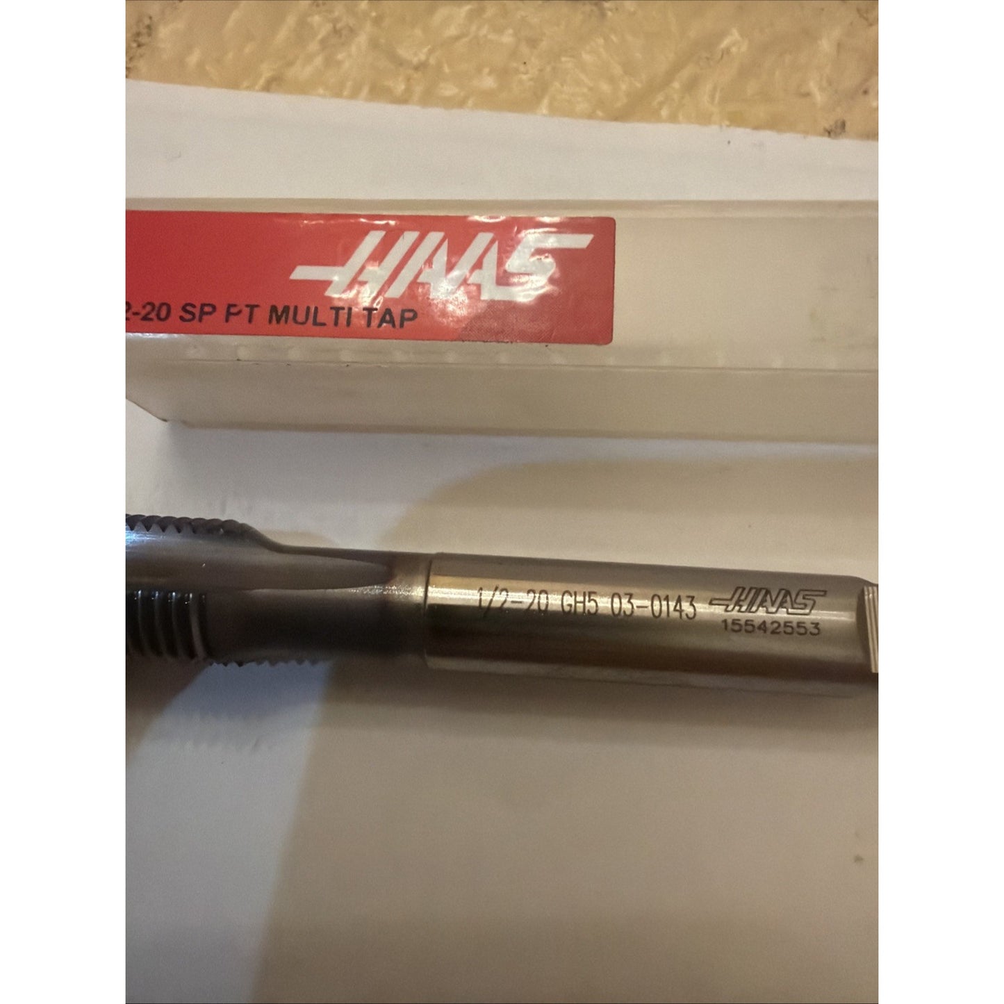 Close-up of a metallic threading tap with engraved size and product numbers along the shank, placed on a white surface next to a clear plastic case with a red Haas label.