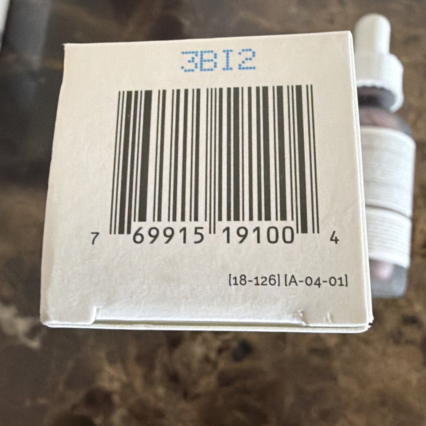 Close-up of the bottom of a white product box showing a barcode with numbers beneath it, a blue printed code above, and additional small printed codes below, with blurred dropper bottles in the background on a textured surface.