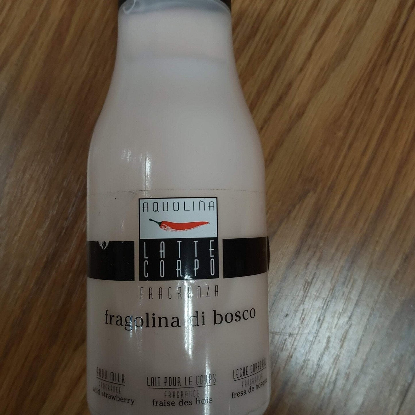 Creamy white bottle of Aquolina Latte Corpo body lotion with woodland strawberry fragrance, featuring a black label strip and red chili pepper logo on a wooden surface.