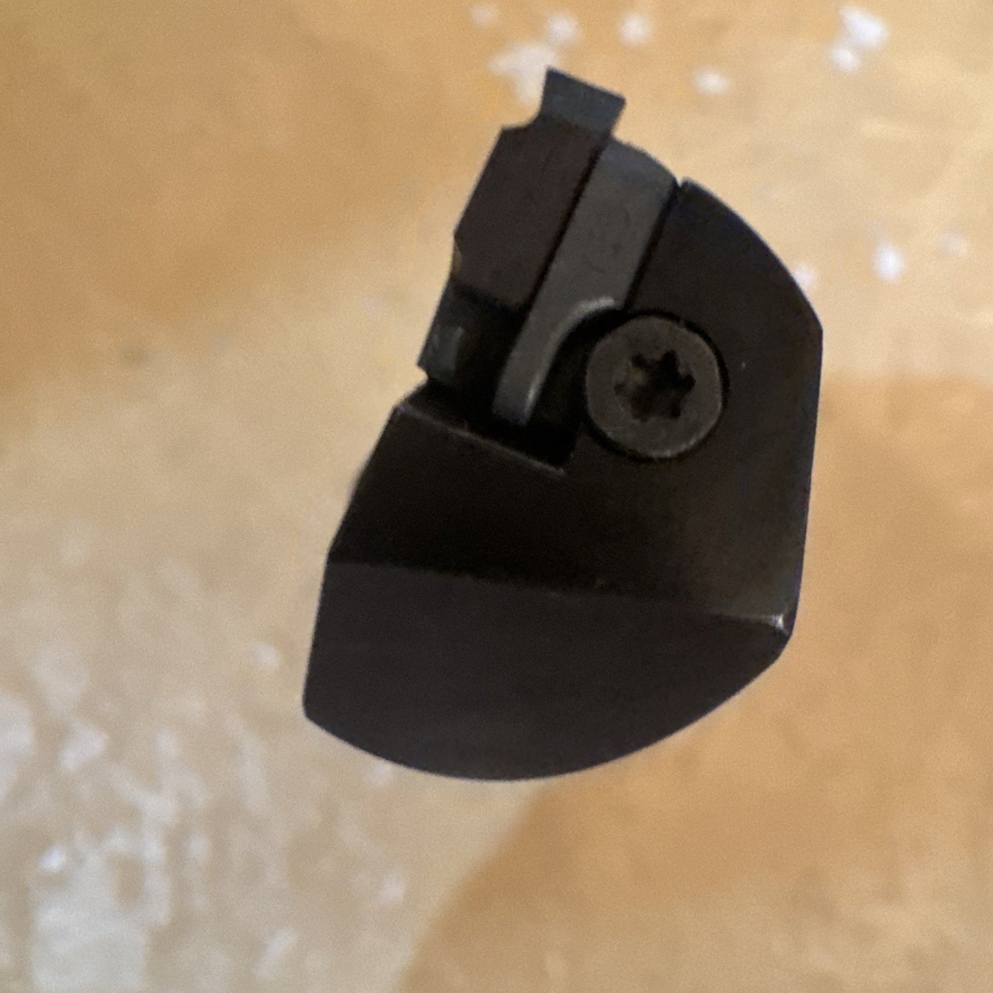 Top-down close-up of a dark metal cutting tool tip with a small rectangular insert secured by a screw, set against a light beige textured background.