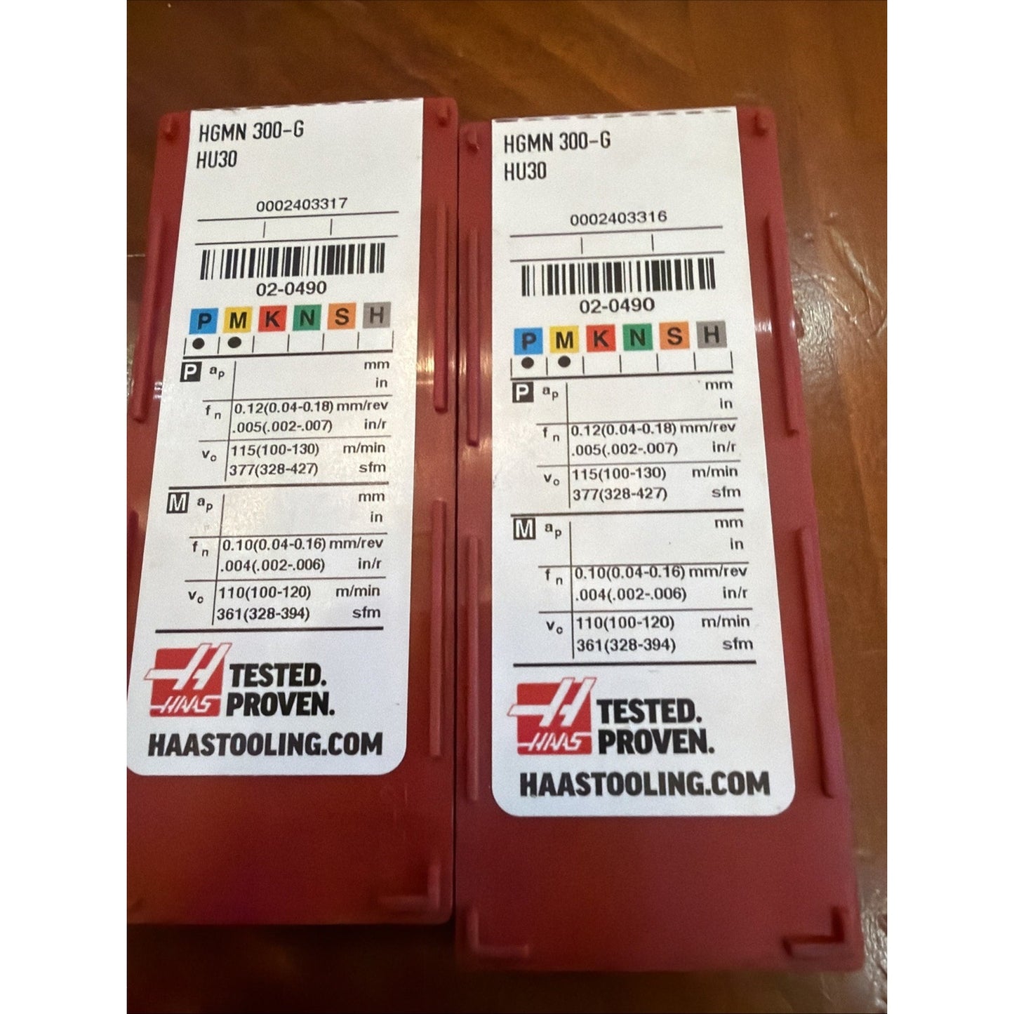 Two red plastic cases with white labels showing product codes "HGMN 300-G HU30," machining parameters in metric and imperial units, ISO classification color blocks, barcodes, and Haas Tested Proven branding on a wooden surface.