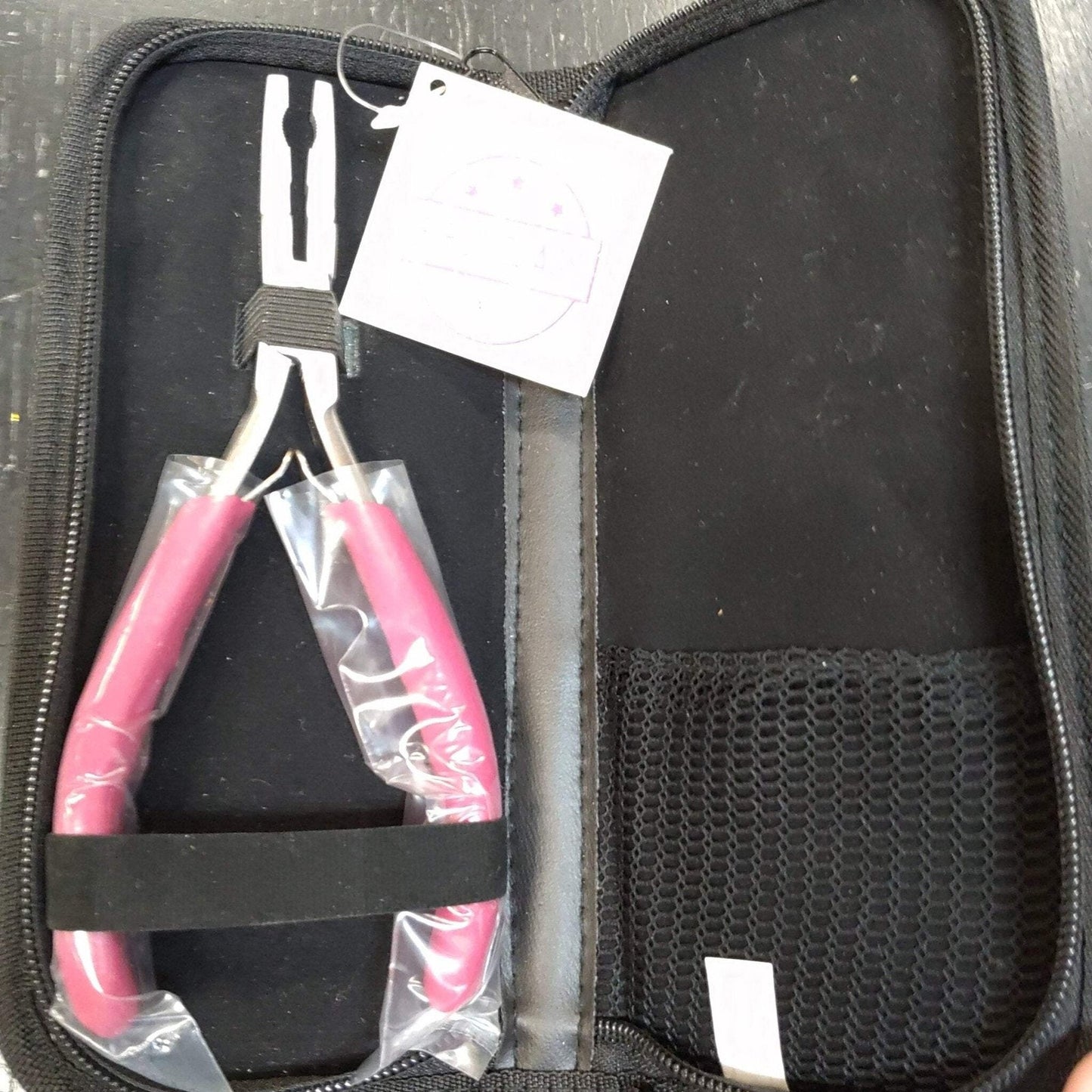Silver pliers with bright pink handles partially wrapped in plastic, secured with a black elastic band inside an open black zippered carrying case with a mesh pocket on the right side.