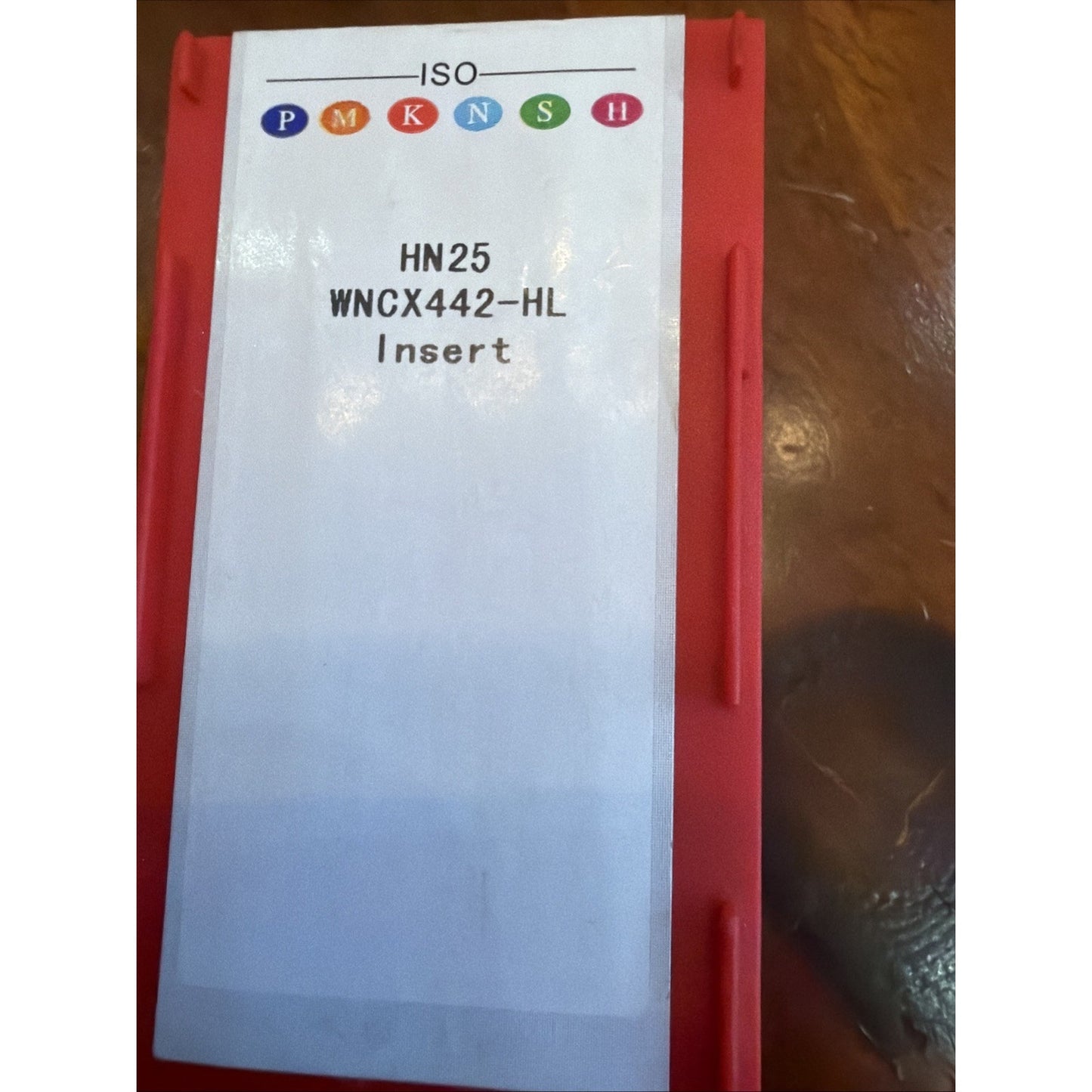Red rectangular container with a white label displaying colored circular ISO symbols and text reading "HN25 WNCX442-HL Insert" placed on a wooden surface.