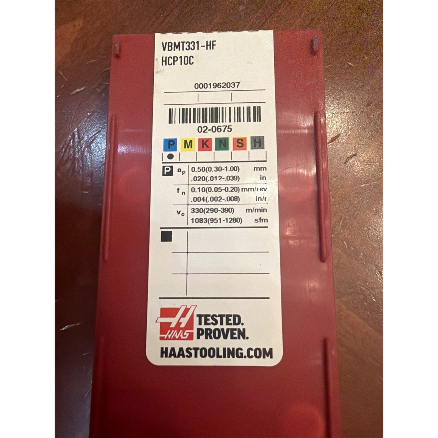 Red plastic rectangular container with a white label showing product code "VBMT331-HF HCP10C," color-coded ISO material classification symbols, machining parameters, barcode, and Haas Tested Proven logo.
