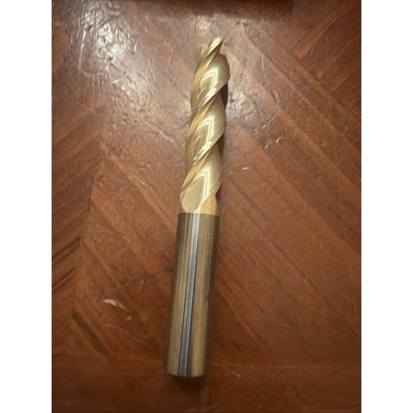 Single metal twist drill bit with a polished shank and sharp, gold-colored flutes resting on a textured wooden surface.