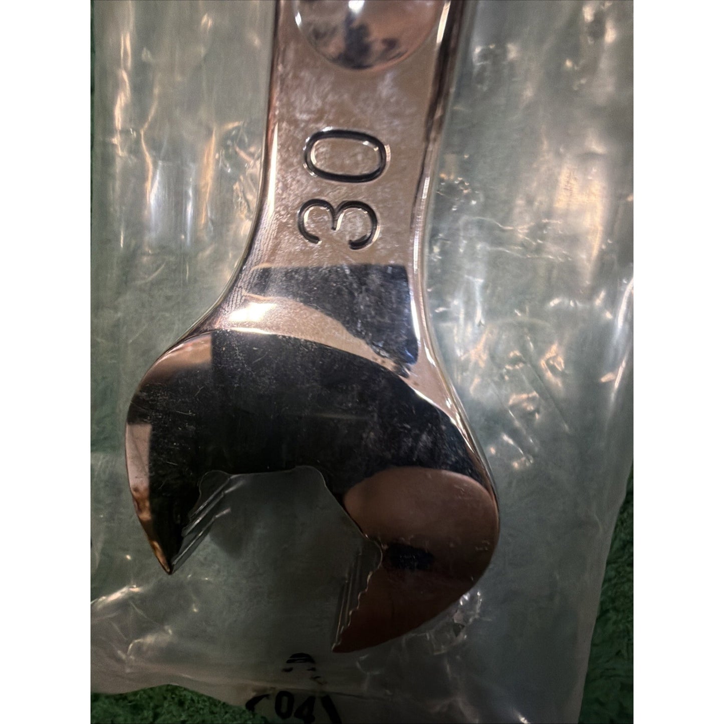 Close-up of a polished metal open-end wrench with the number 30 engraved, resting on a translucent plastic surface over a textured green background.