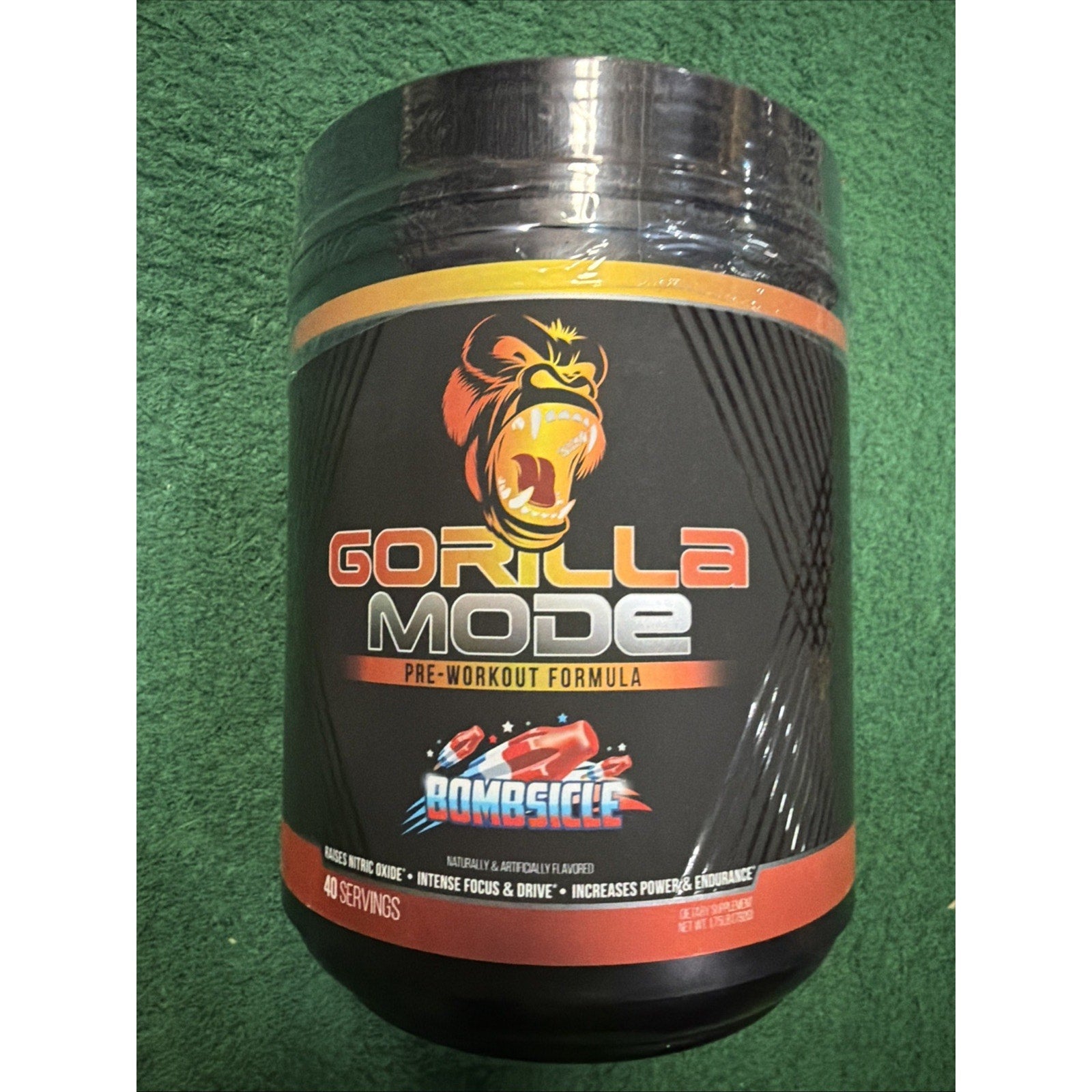 Black supplement container with an orange and yellow accented label featuring an illustrated roaring gorilla, titled "Gorilla Mode" with flavor "Bombsicle," placed on green textured fabric.