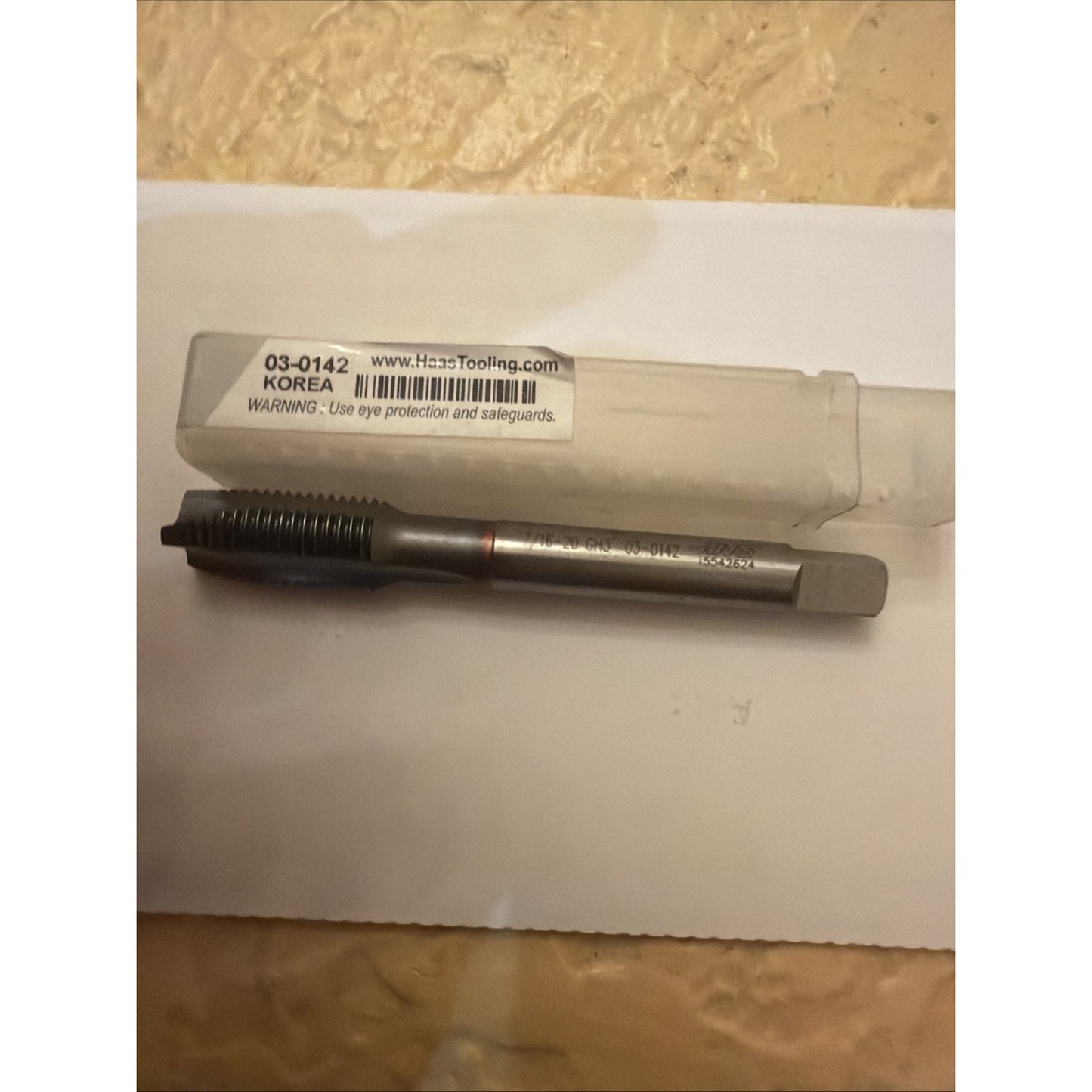 Metal threading tap with a square shank and straight flutes, resting on a white surface next to a clear plastic storage tube labeled with product code and safety warning.
