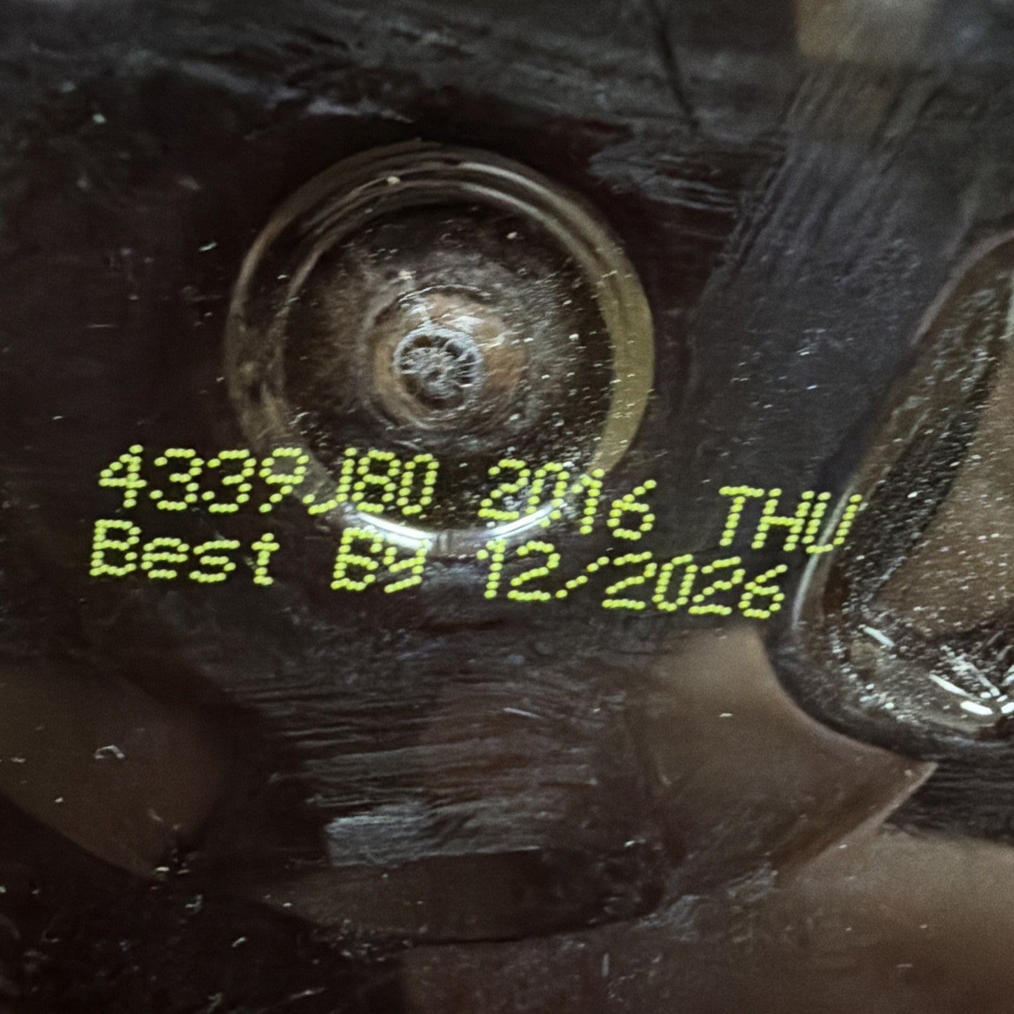 Close-up of yellow printed code "4339.180 2016 THU" and best by date "12/2026" on a dark textured surface under glossy plastic.