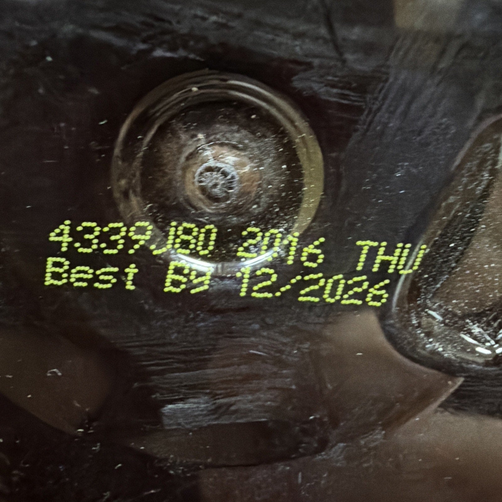 Close-up of yellow printed code "4339.180 2016 THU" and best by date "12/2026" on a dark textured surface under glossy plastic.
