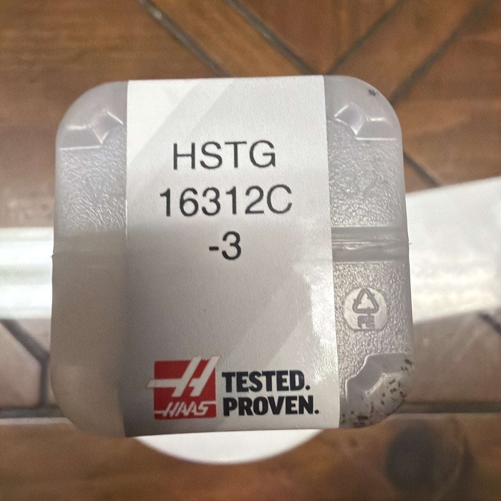 Small translucent plastic container with a white label displaying the code "HSTG 16312C -3" and a red and black "Tested. Proven." logo on a wooden surface.
