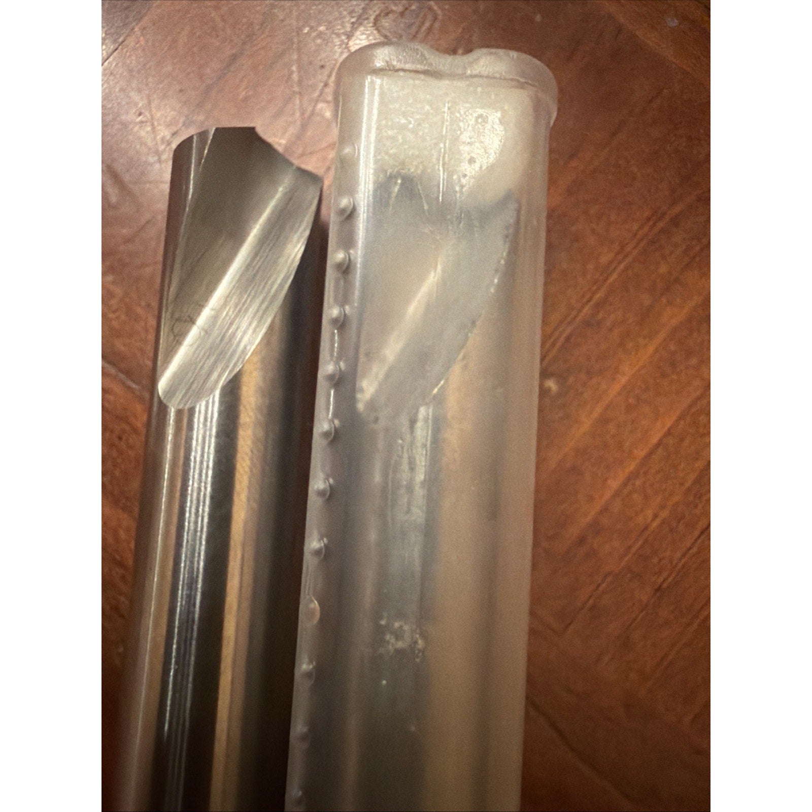 Close-up of a shiny metal cutting tool with a spiral flute alongside a translucent plastic case on a wooden surface.
